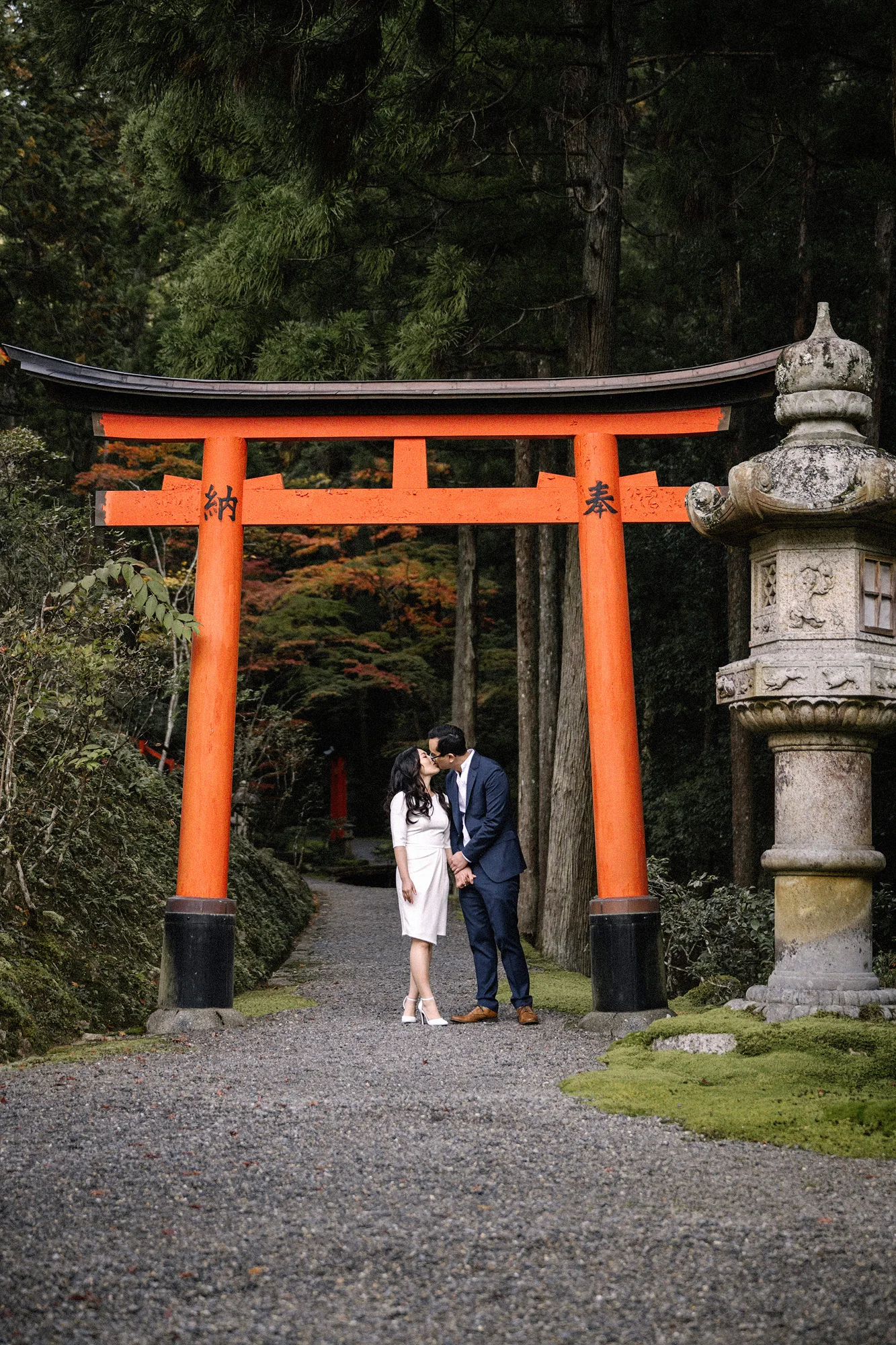 A couple stand together beneath a vermilion torii gate in a Kyoto cedar forest.