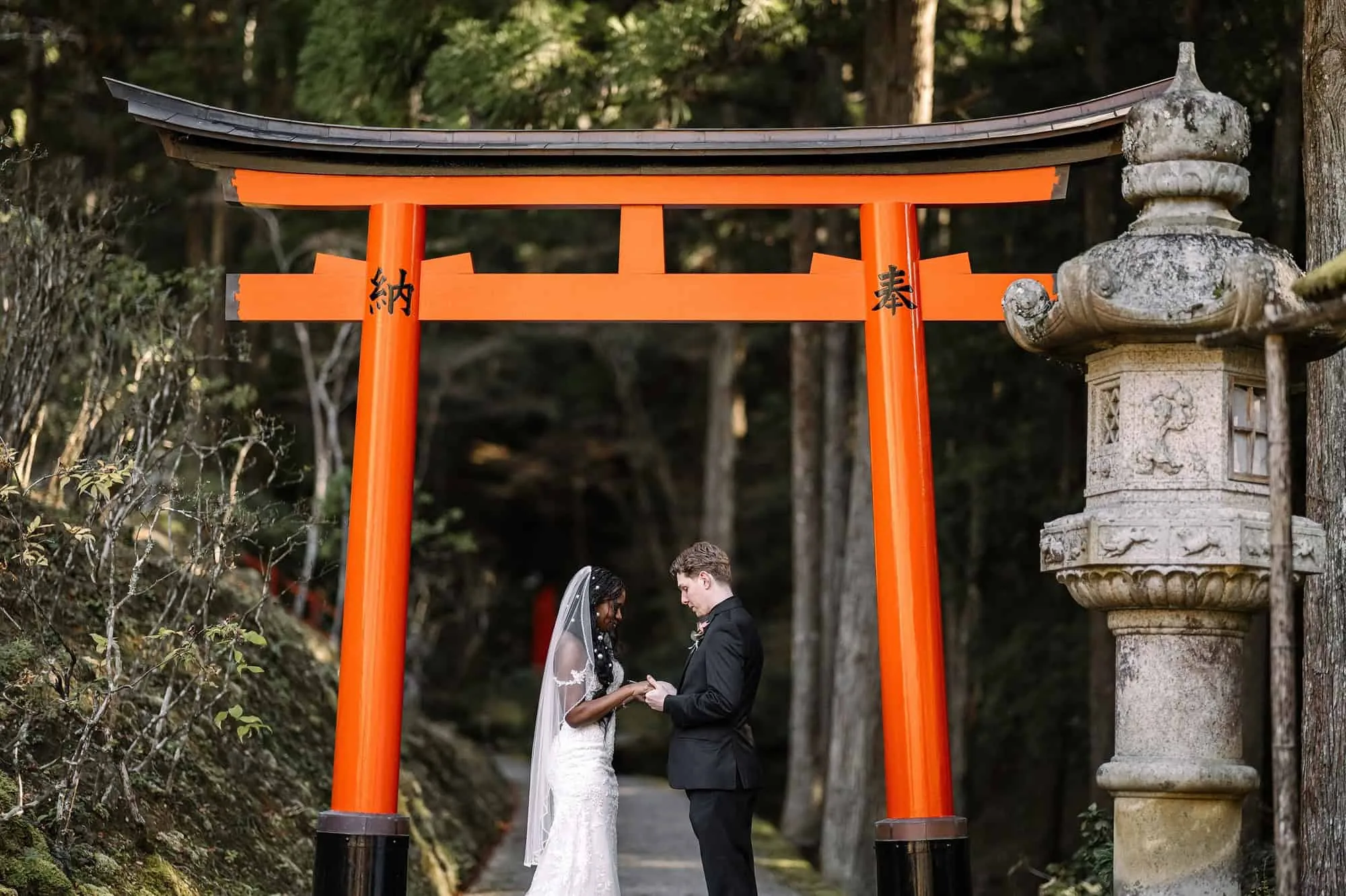 The bride and groom stand close beneath a vermilion torii beside a tall stone lantern in a cedar forest at their Kyoto elopement.