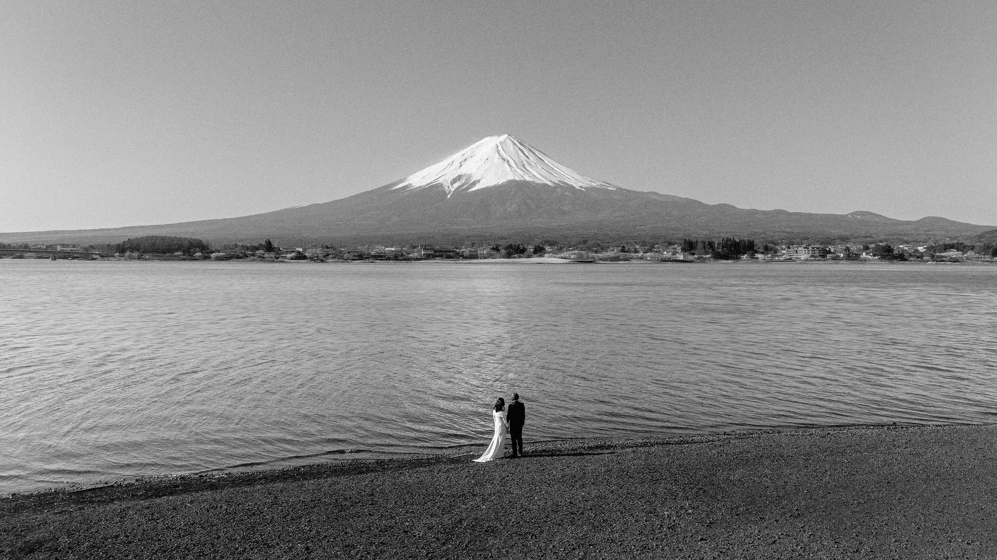 In black and white, a couple stand small at the edge of Lake Kawaguchiko, Mt Fuji across the water.