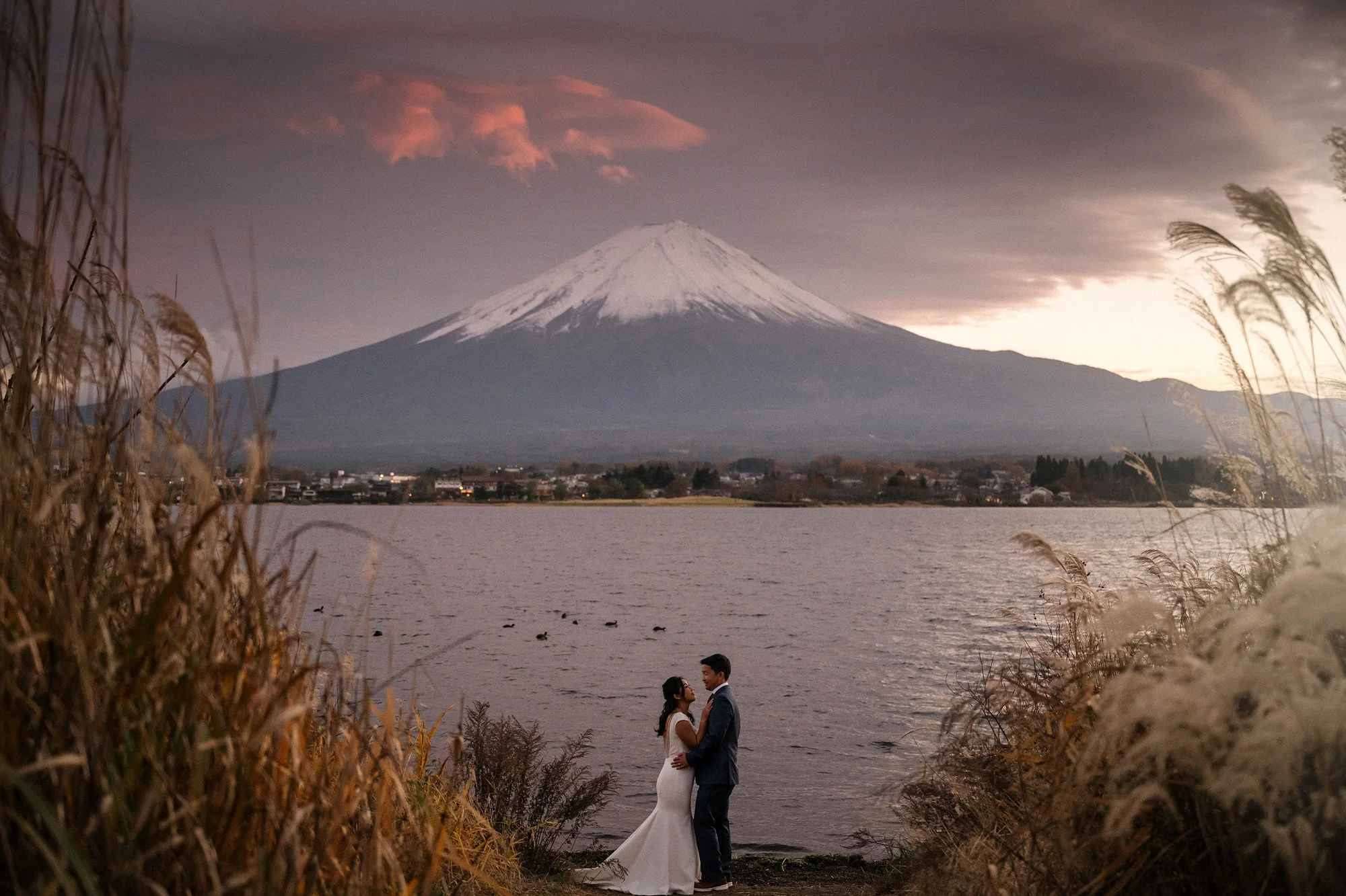 A couple stand at the edge of Lake Kawaguchiko at sunset, Mt Fuji across the water and pampas grass around them.