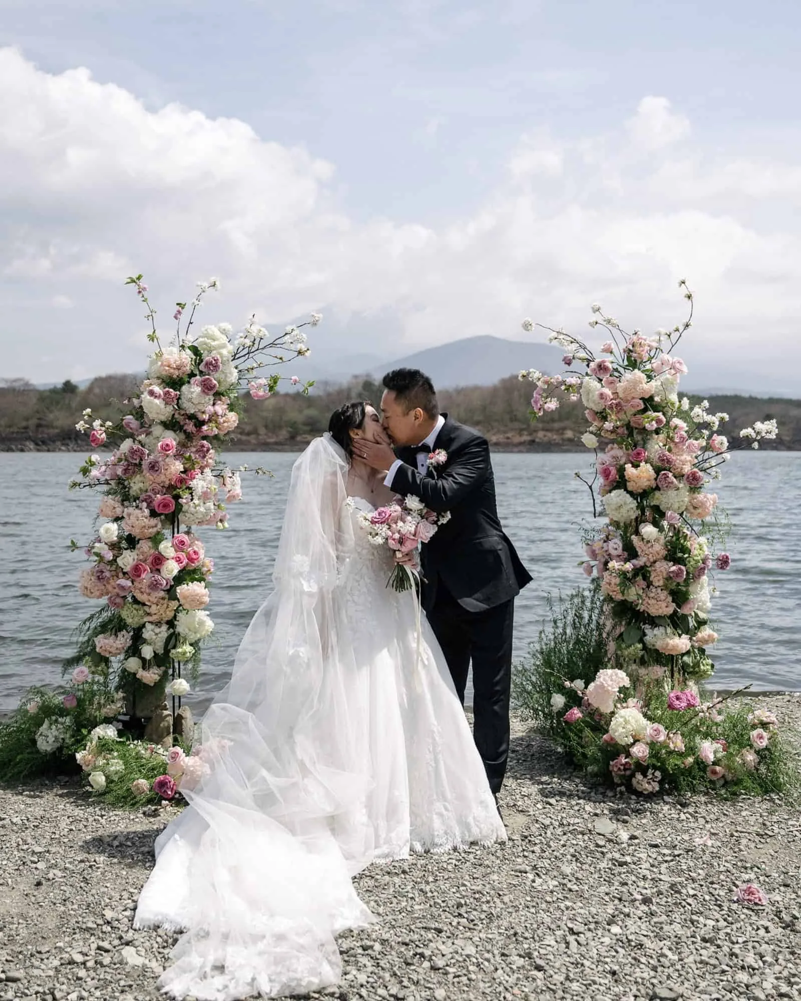 The bride and groom share their first kiss between two floral columns at a Lake Shoji wedding ceremony, the water and a mountain behind them.