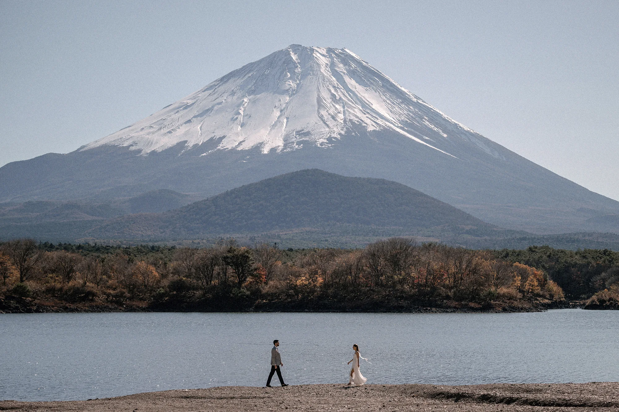 A couple walk the shore of Lake Shoji, Mt Fuji rising above the autumn forest across the water.
