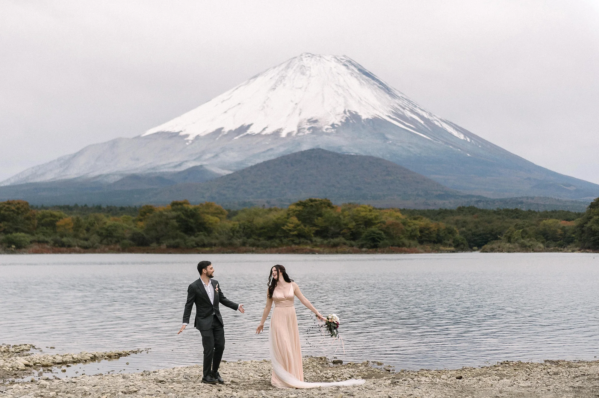 A couple walk the shore of Lake Shoji, the bride in a pale pink gown, Mt Fuji across the water.