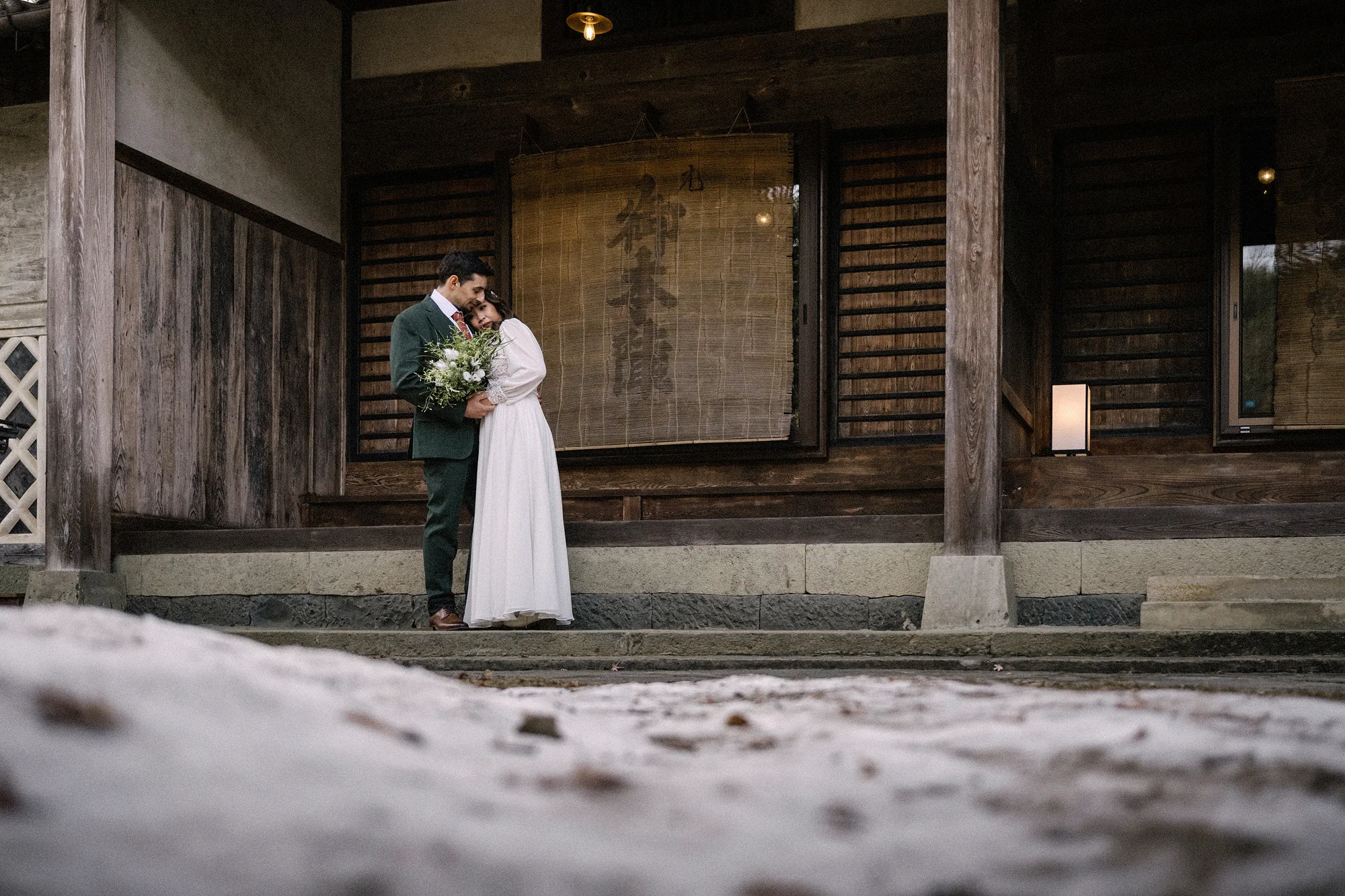 The couple embrace on the veranda of an old timber house, snow on the ground before them, near Matsumoto.