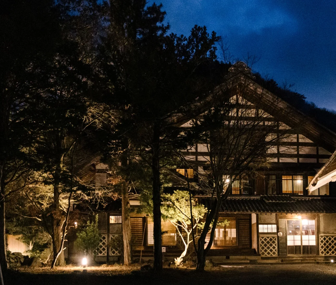 An old timber villa glows with warm light at blue hour, dark pines around it, near Matsumoto.