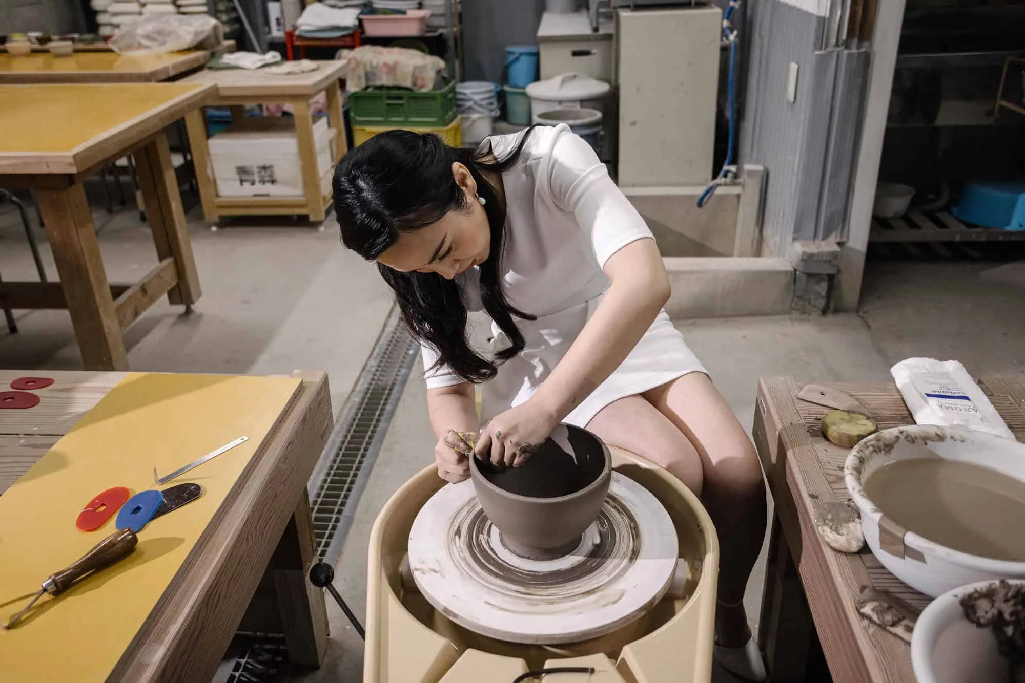 The bride in a short white dress leans over the wheel to shape a clay bowl, tools laid out on the bench, in a pottery studio in Mie.