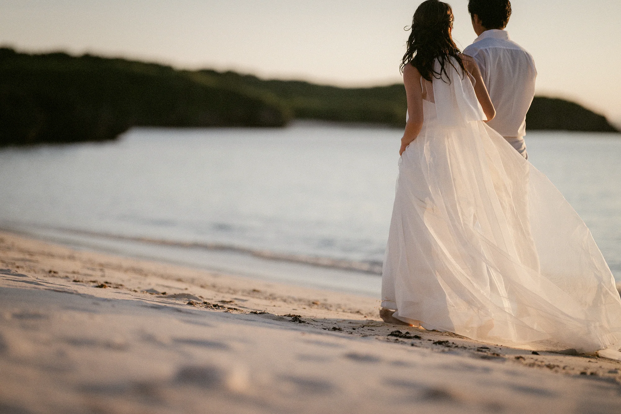 The bride and groom walk along a quiet Miyakojima beach, her gown lifting in the breeze.