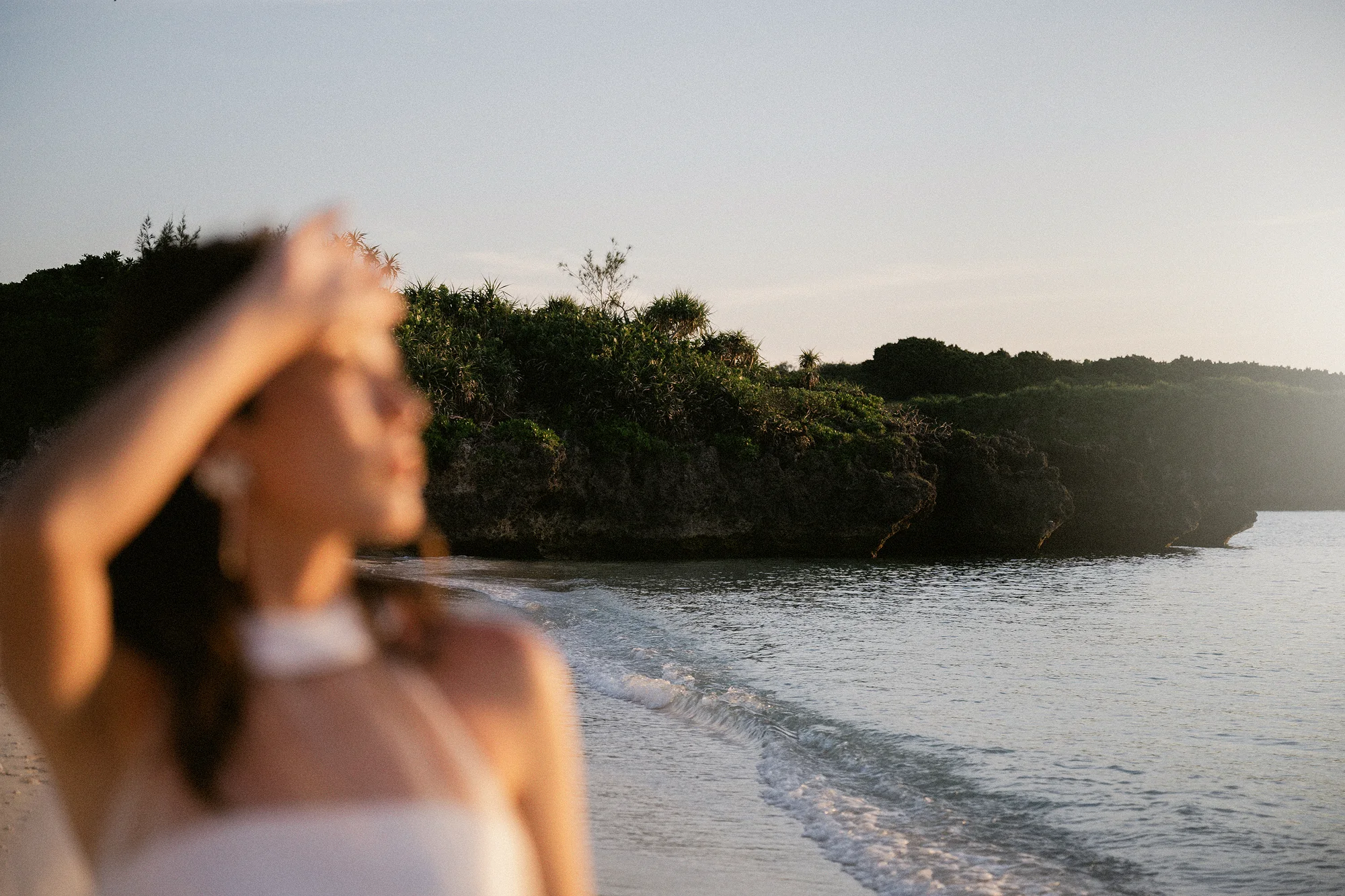 The bride holds back her hair in the sea wind on a Miyakojima beach, a green headland across the water.