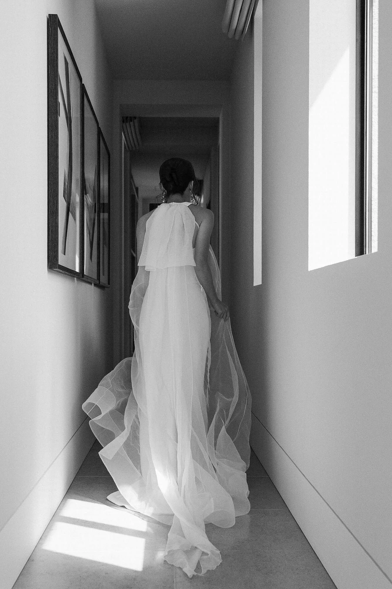 In black and white, the bride walks away down a narrow corridor lined with framed art, her sheer train trailing, on her Miyakojima wedding morning.