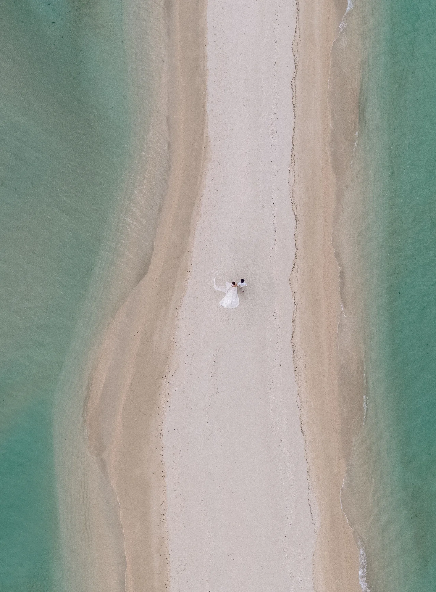 From above, a couple stand on a narrow white sandbar between turquoise shallows off Miyakojima.