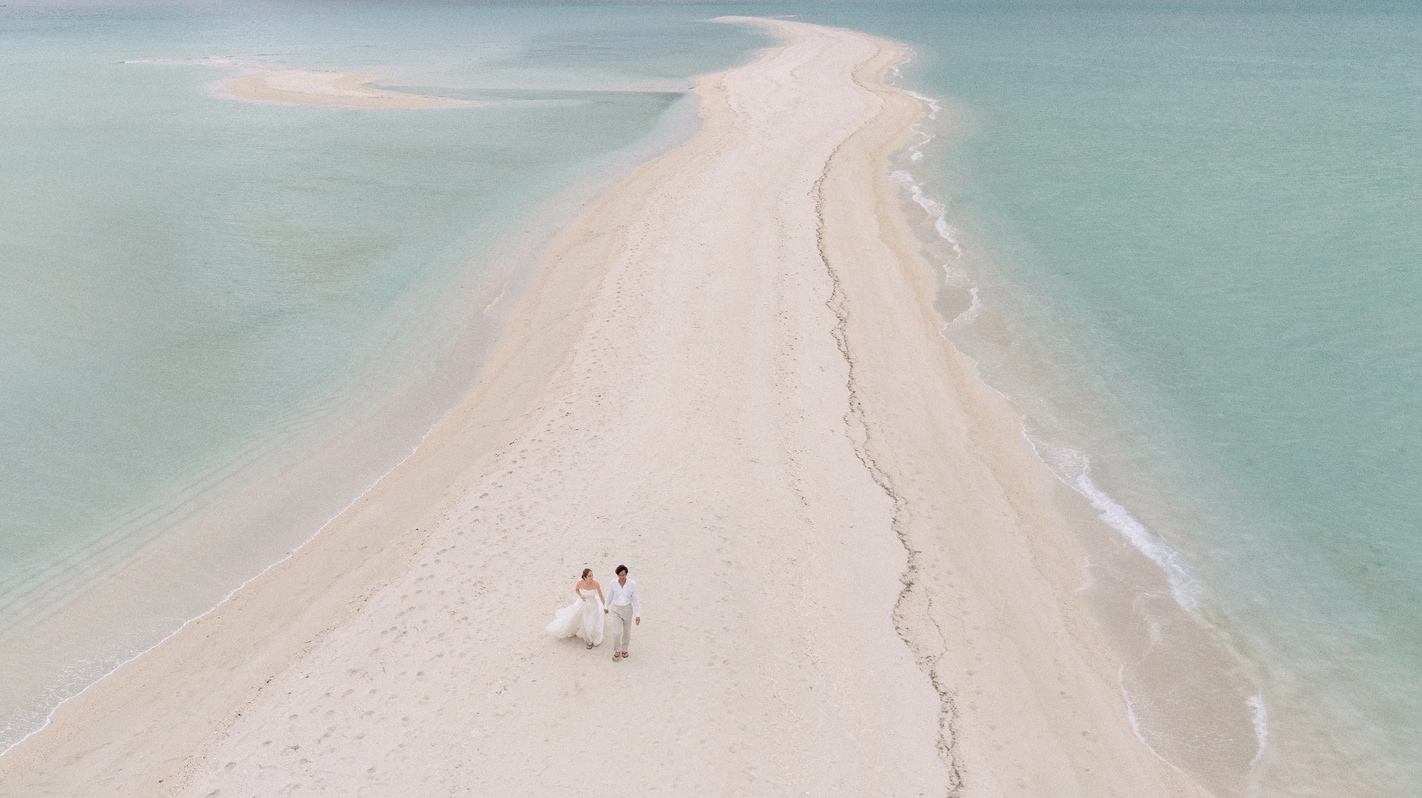 A long white sandbar curves into turquoise water, the couple small at its center.