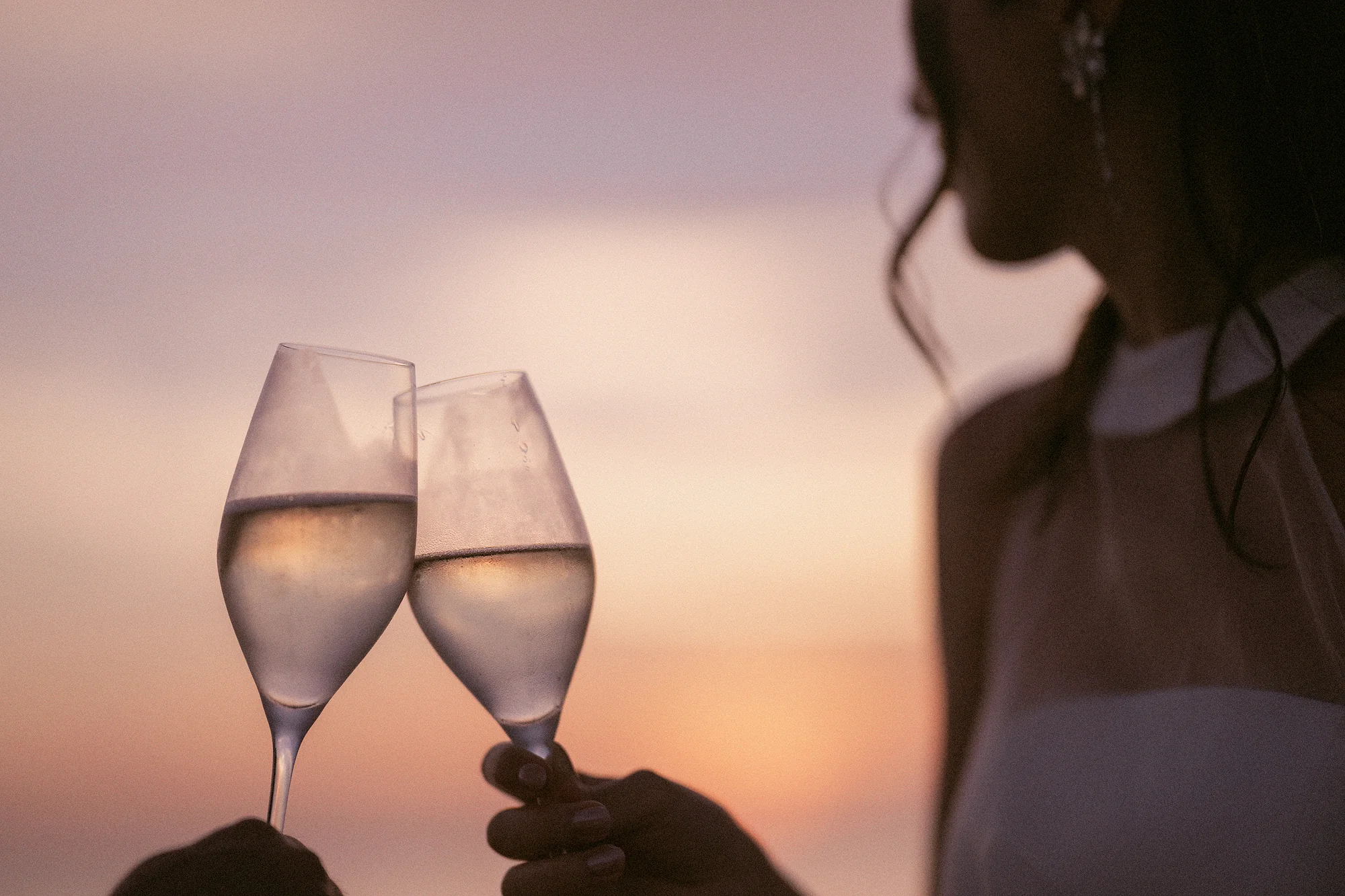 Two glasses meet against a pink Miyakojima sunset, the bride in soft focus behind.
