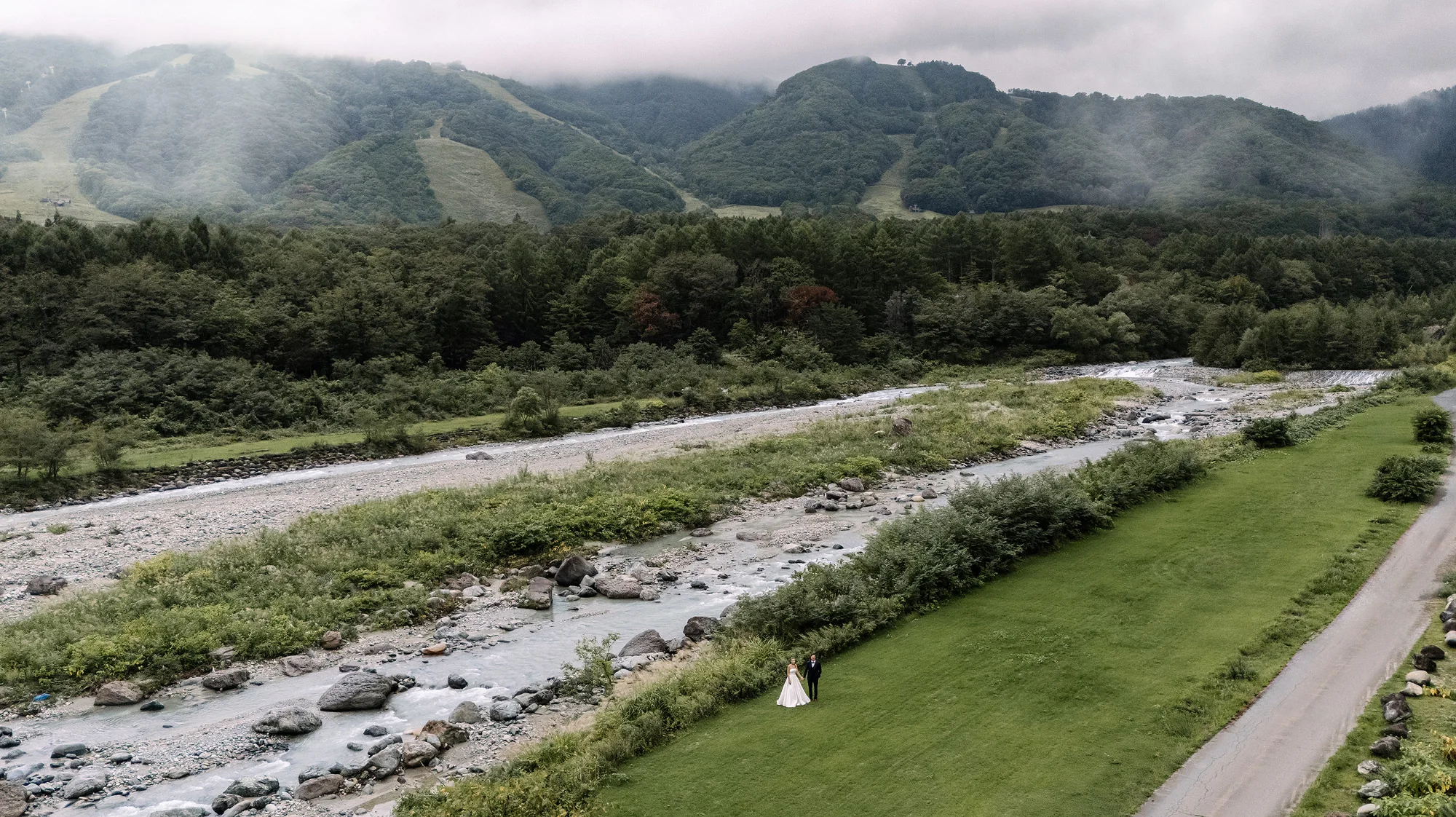 From above, a couple stand on a green riverbank in a Hakuba valley in summer, mist on the ski slopes behind.
