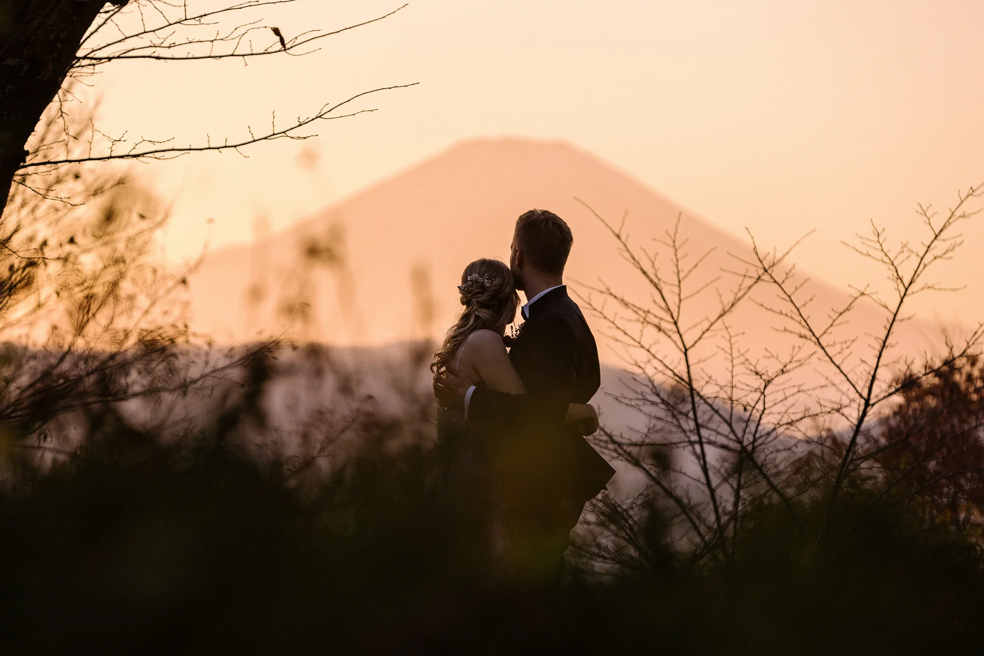 The couple embrace in silhouette as Mt Fuji glows pink and gold at dusk behind them.