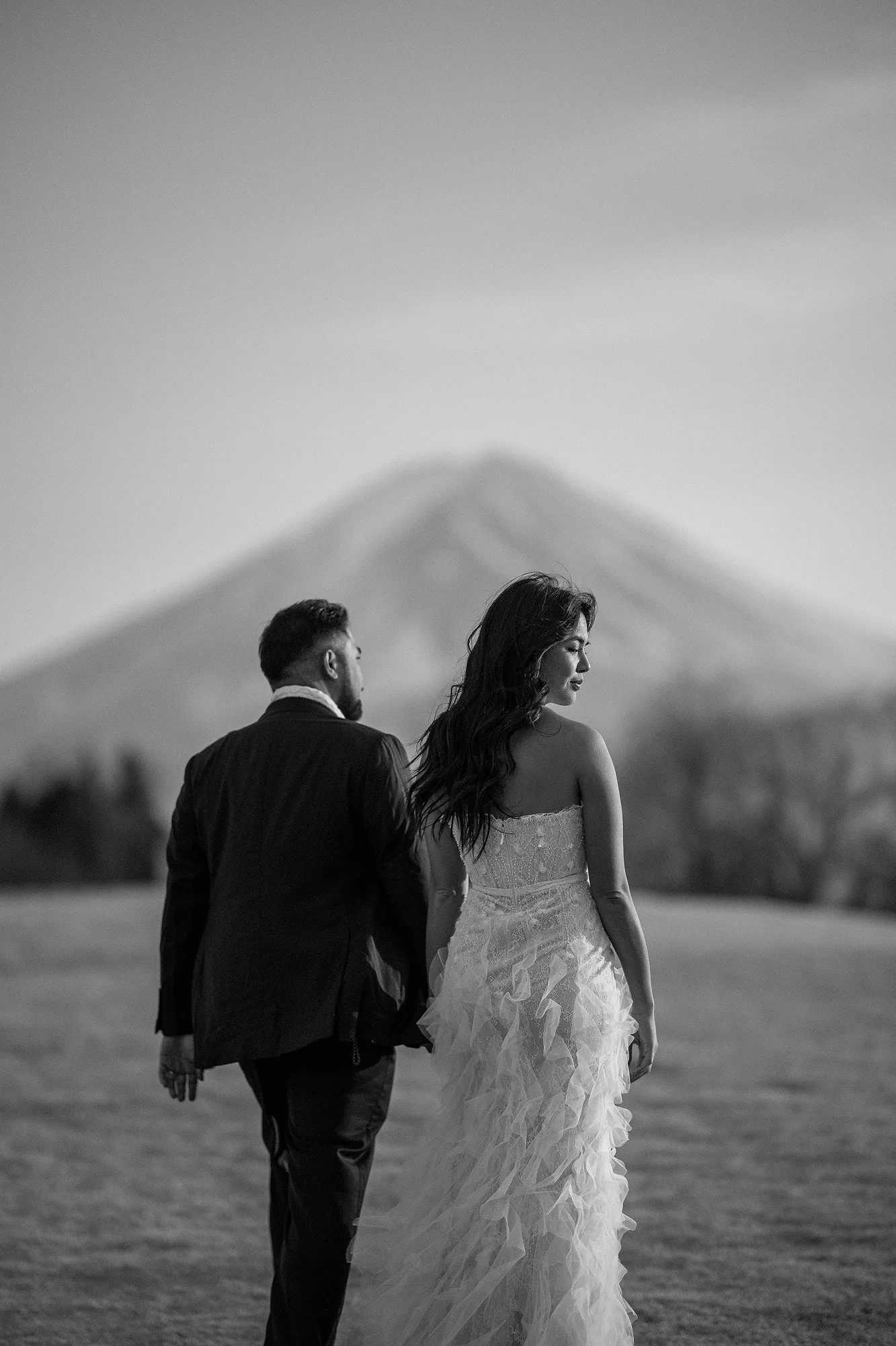 In black and white, a couple walk hand in hand toward Mt Fuji.