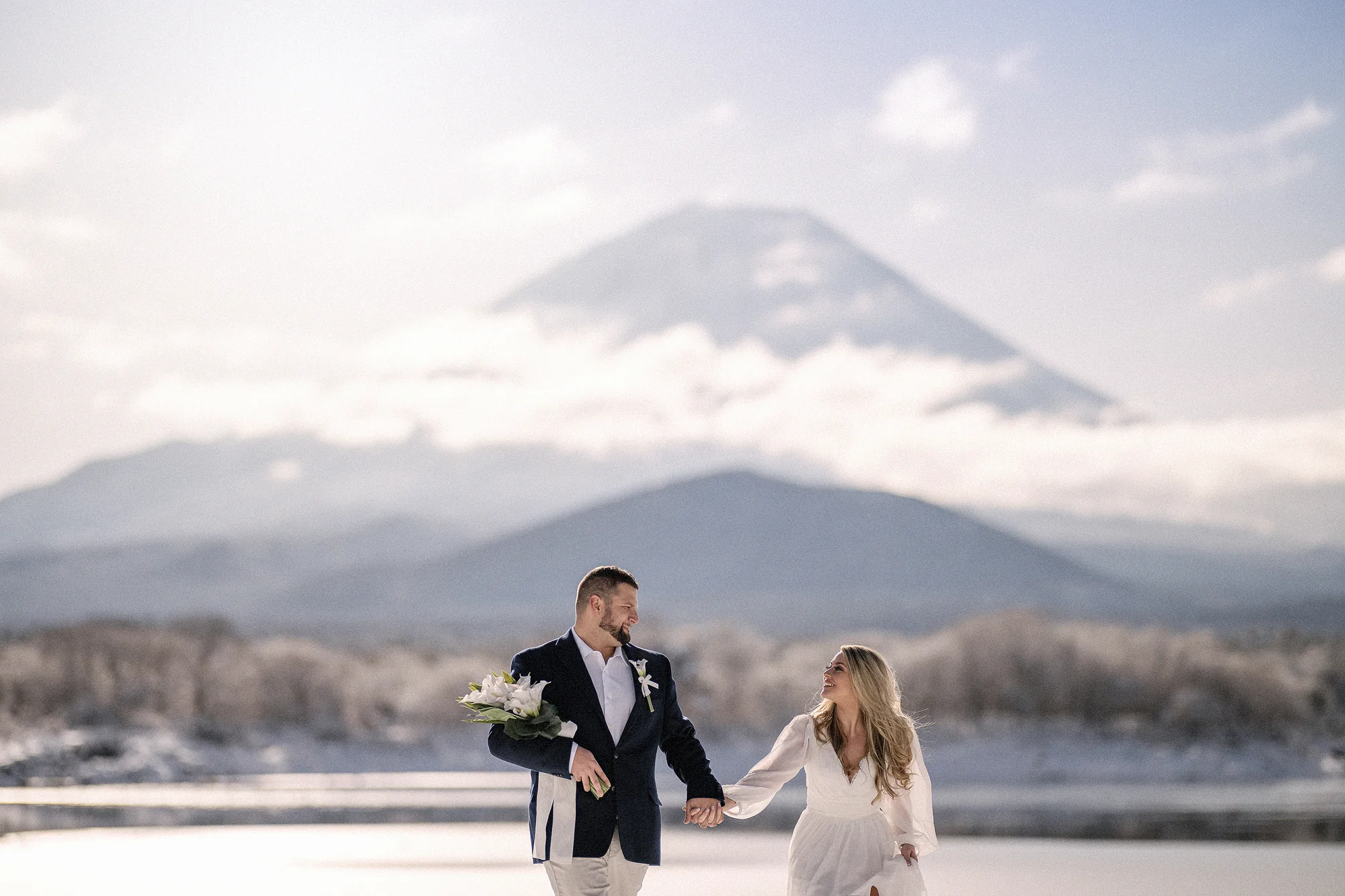 The couple walk hand in hand along a frosted lakeshore, Mt Fuji rising through soft cloud behind them.