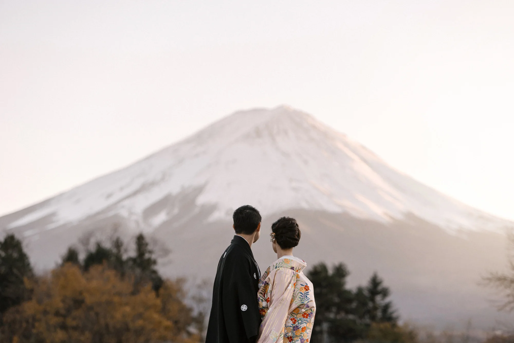 A couple in kimono stand together at dusk, snow-capped Mt Fuji filling the sky behind them.