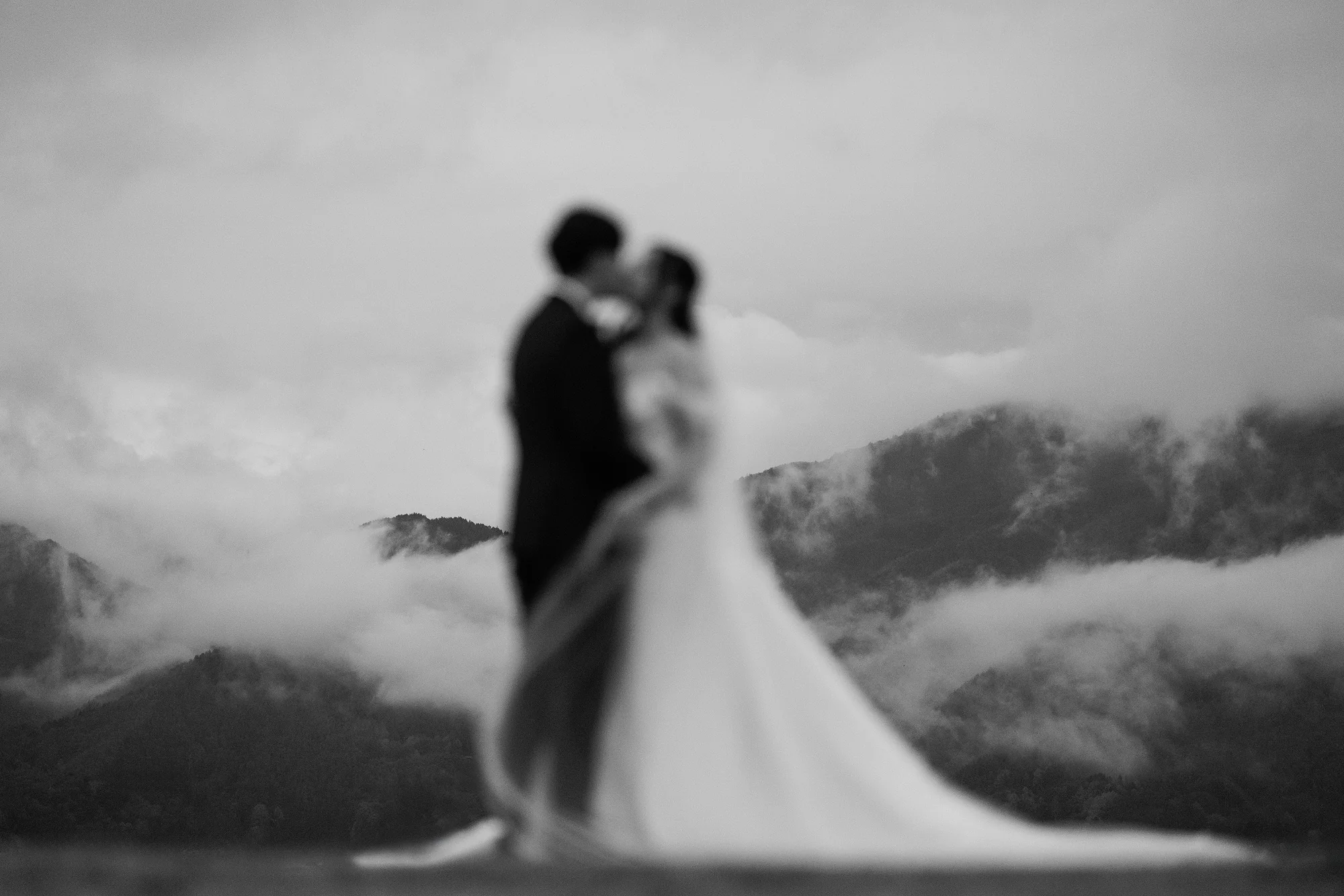 In black and white, a couple kiss on a hillside as cloud fills the valley below Mt Fuji.