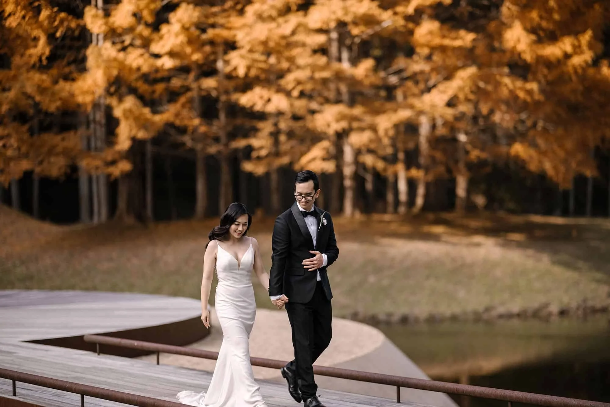 The couple walk along a pond edge beneath golden autumn larches at the art forest in Nara.