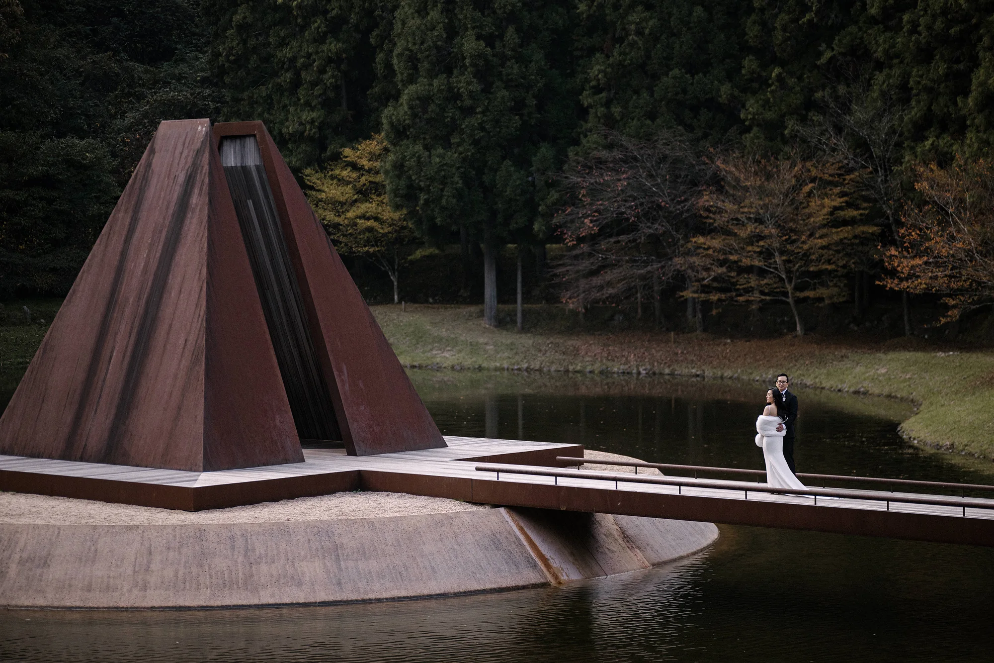 The bride stands at the edge of a pond beside a rusted steel pyramid at the art forest in Nara.