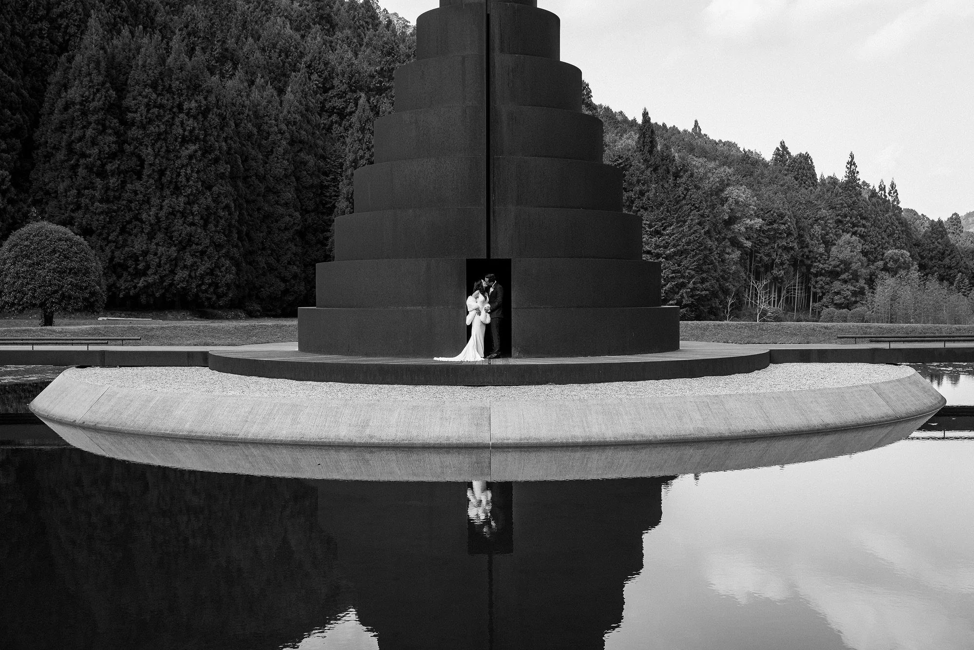 In black and white, a couple stand before a stepped black tower reflected in still water at Murou Art Forest.