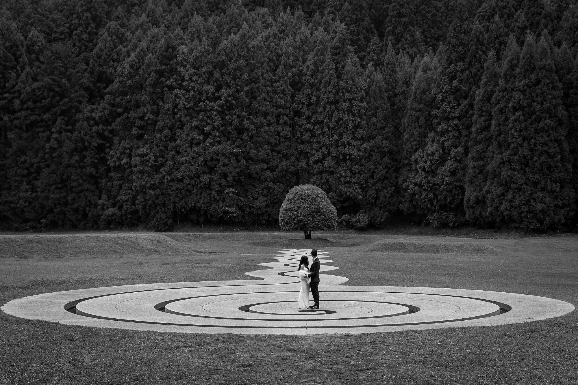 In black and white, a couple stand in the stone spiral at Murou Art Forest, a single round tree beyond.