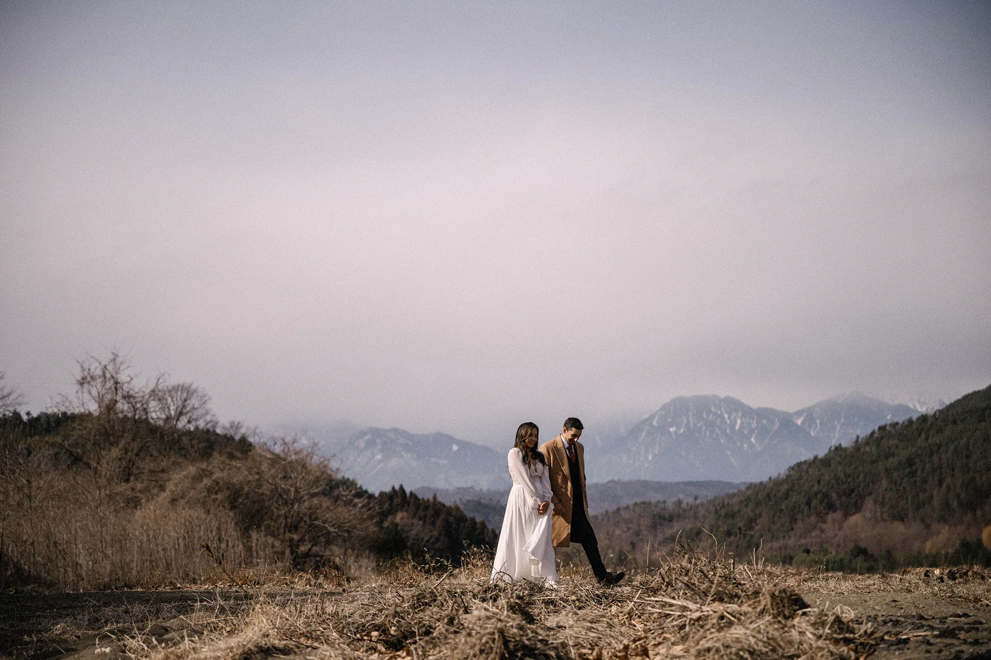 The couple walk across a winter hilltop, the snowy peaks of the Japanese Alps behind them, in Nagano.