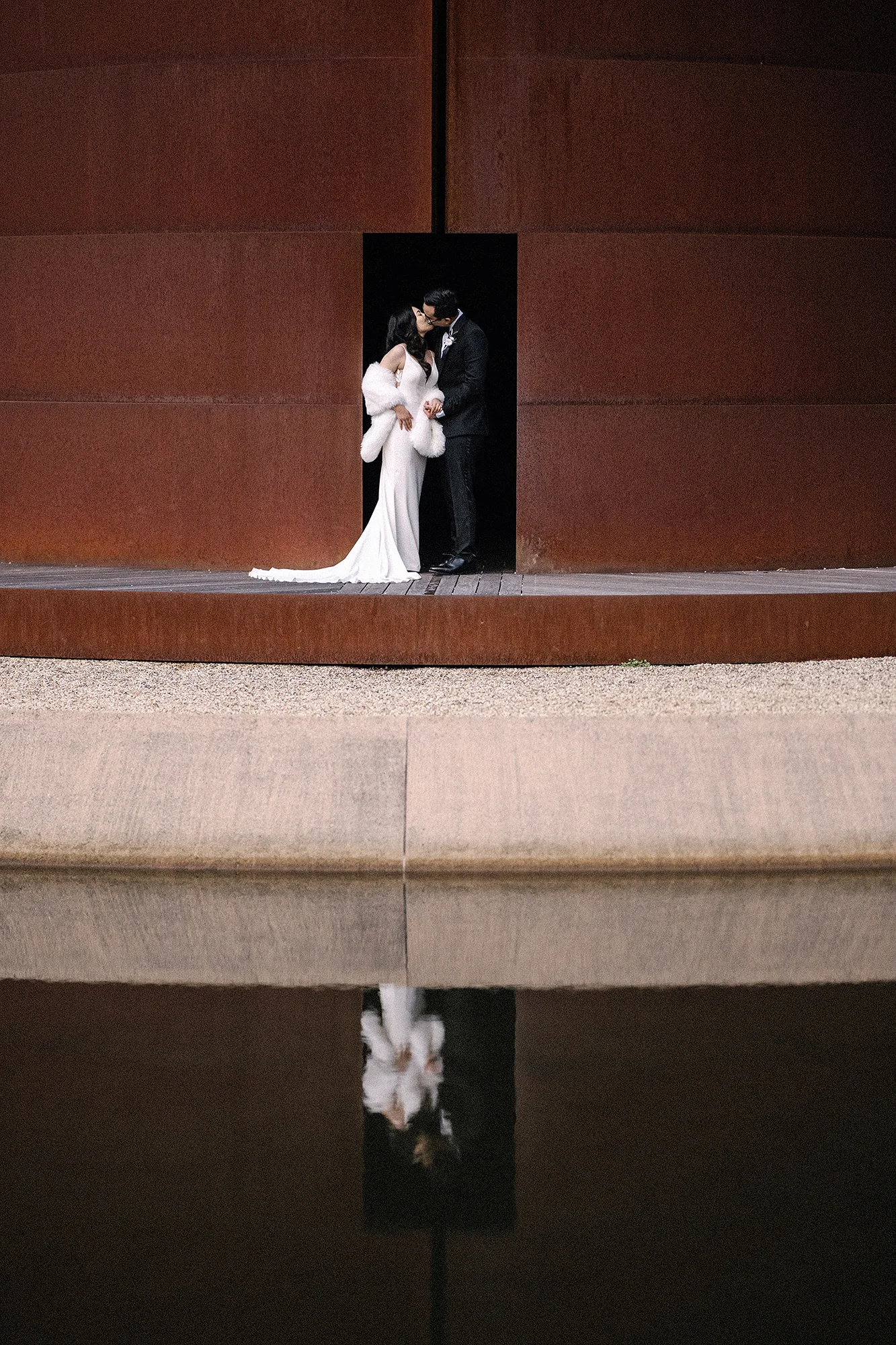 The couple stand in the doorway of a rusted steel sculpture, reflected in still water at an art forest in Nara.