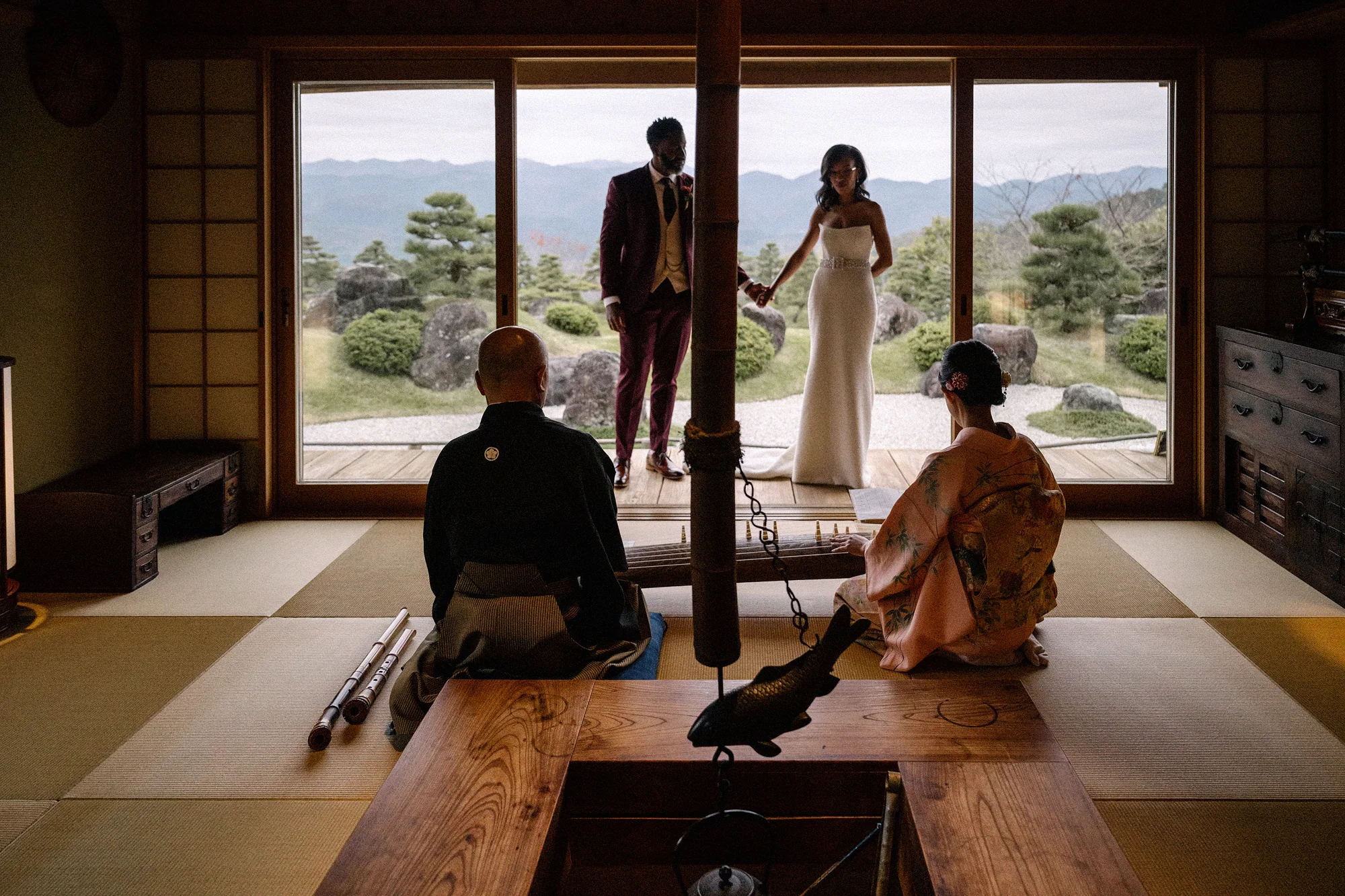 The couple stand at the open window of a tatami room with a hearth, the mountains beyond, as musicians play nearby.