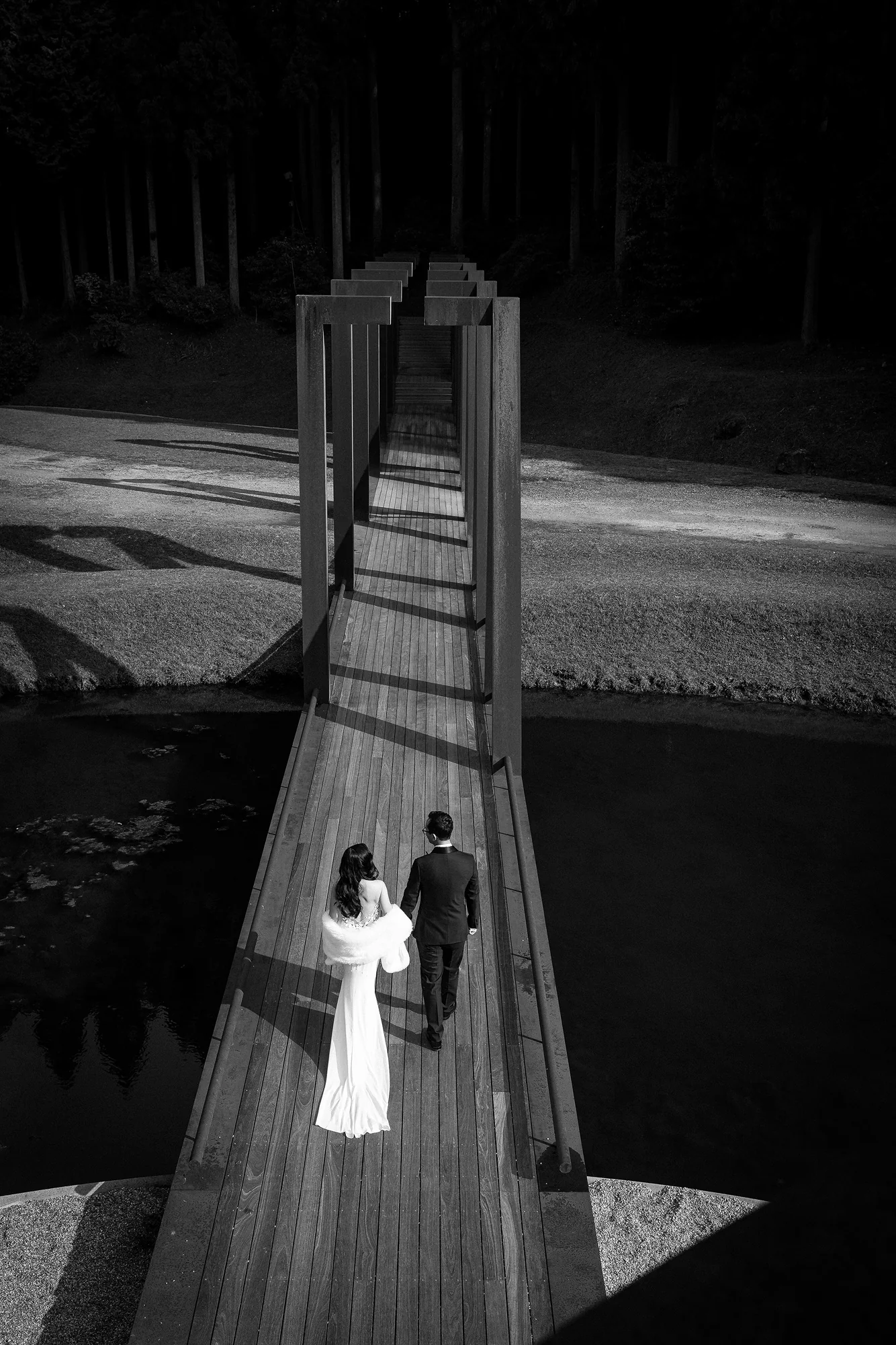 In black and white, a couple walk out along a narrow wooden bridge over a dark pond.