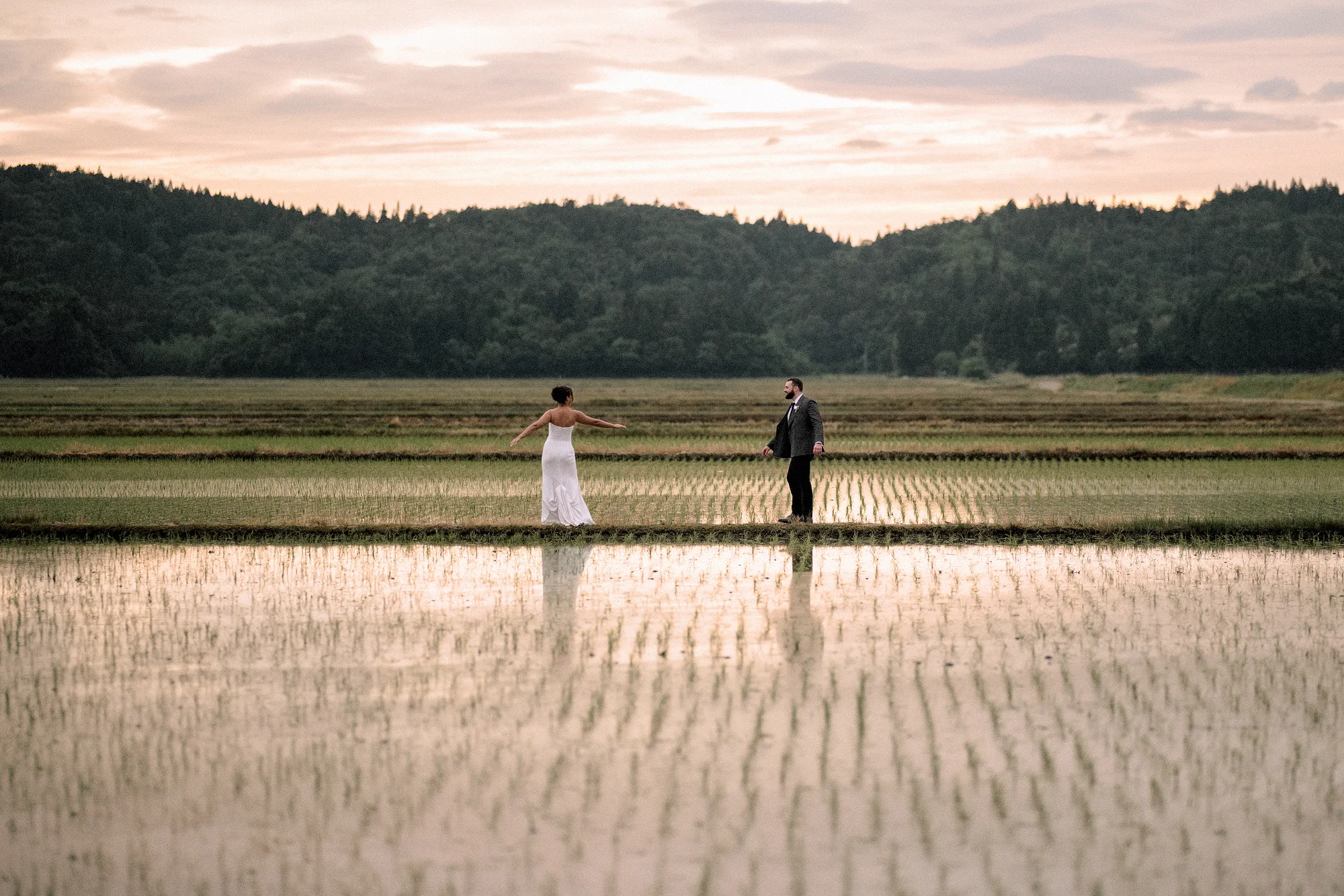 The couple walk through flooded rice paddies at dusk in Niigata, the evening sky reflected around them.