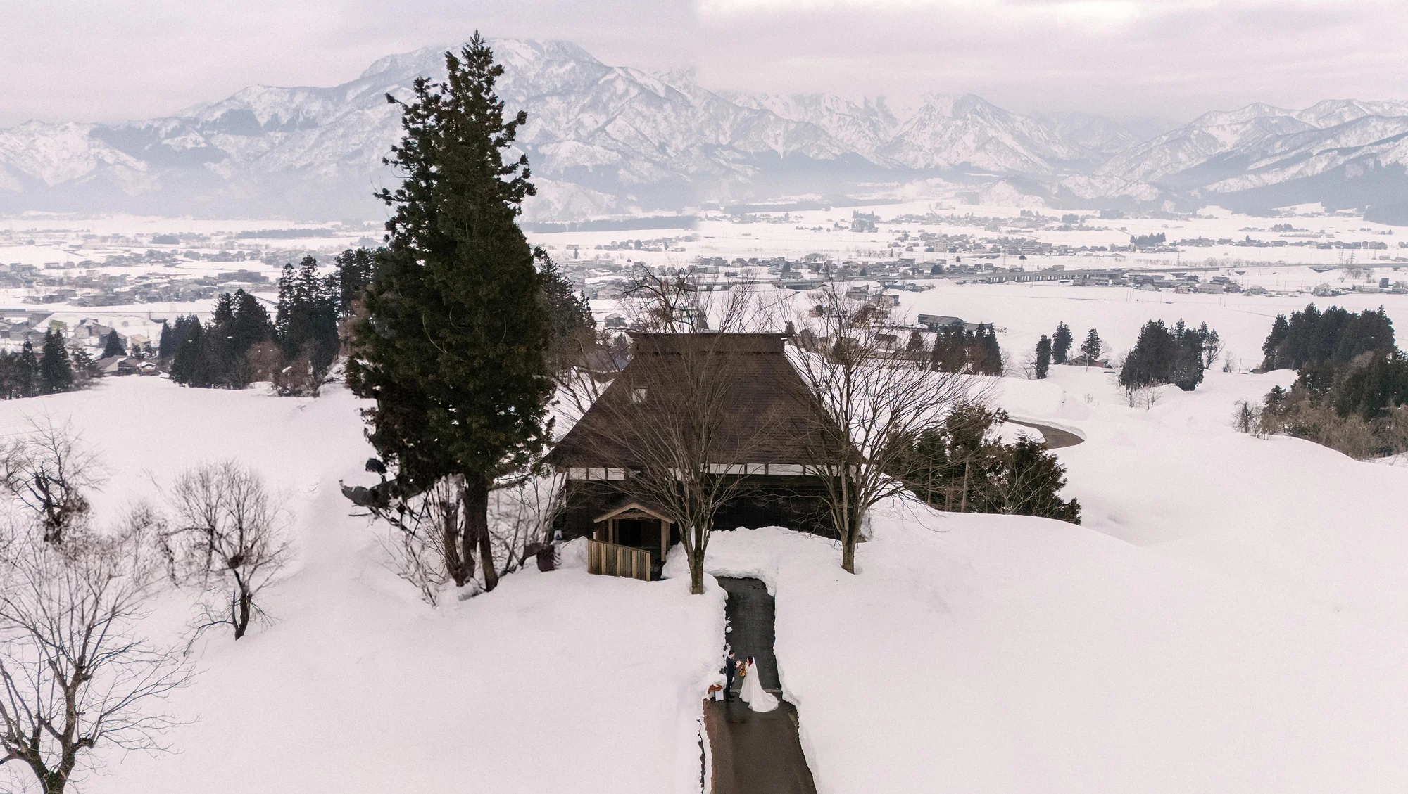 A couple walk a snowy path toward a thatched farmhouse, mountains beyond.