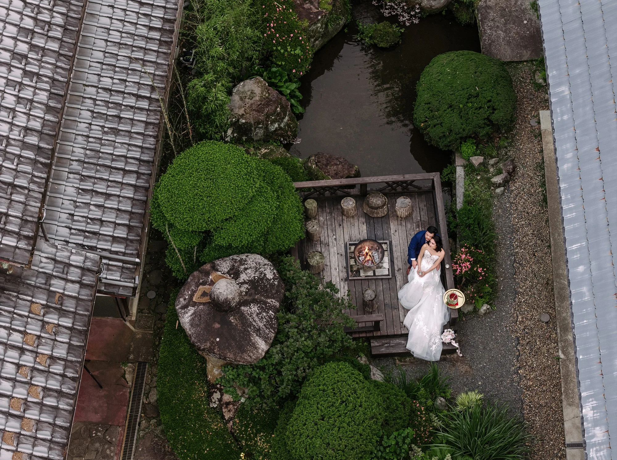 From above, the couple sit by a fire pit in a small courtyard garden beside old tiled roofs at NIPPONIA Kosuge.