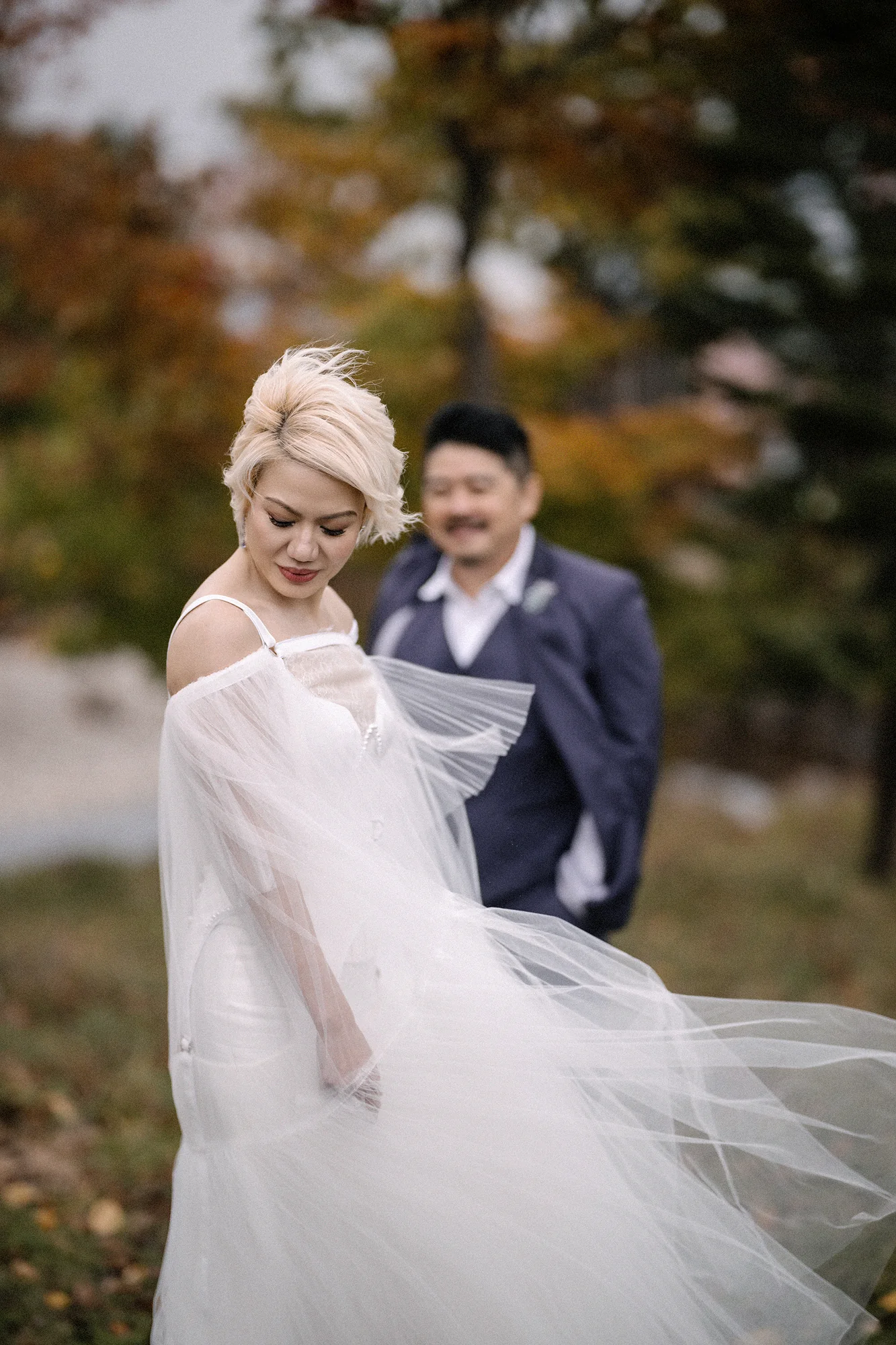 The bride's sheer cape lifts in the wind as the groom looks on, autumn trees behind them in Niseko.