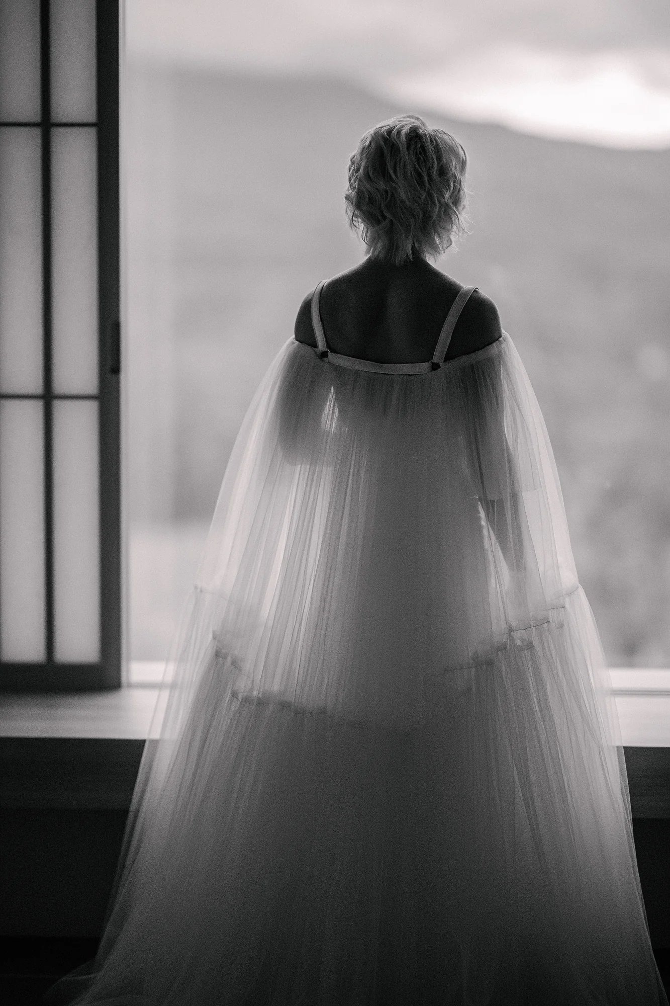 In black and white, the bride stands at a tall window in her sheer cape, the hills outside soft with cloud.