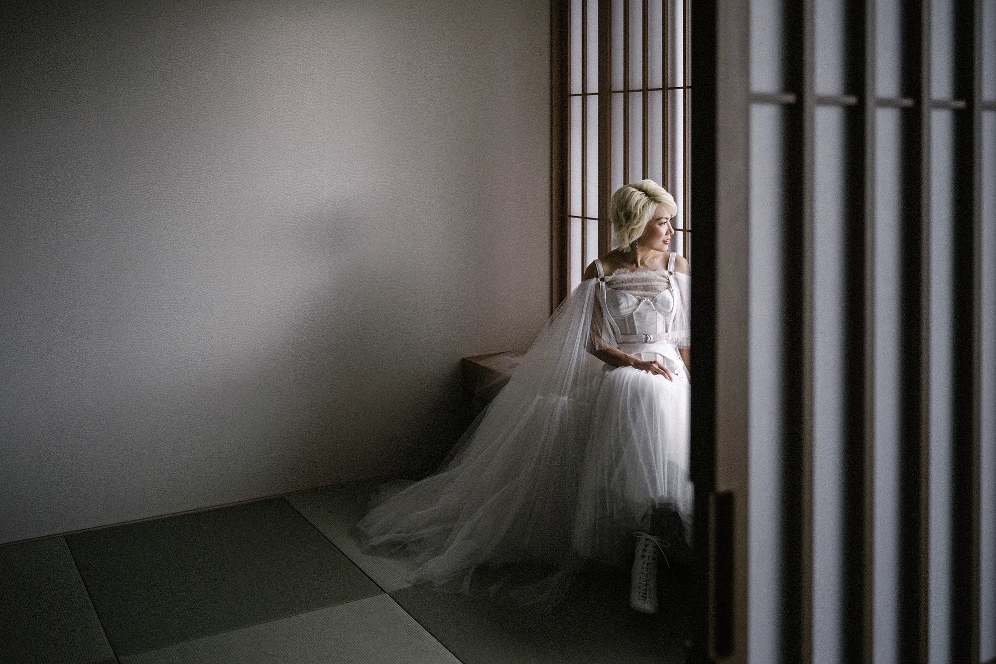 The bride sits by a slatted window in a quiet tatami room, soft light across her gown, in Niseko.