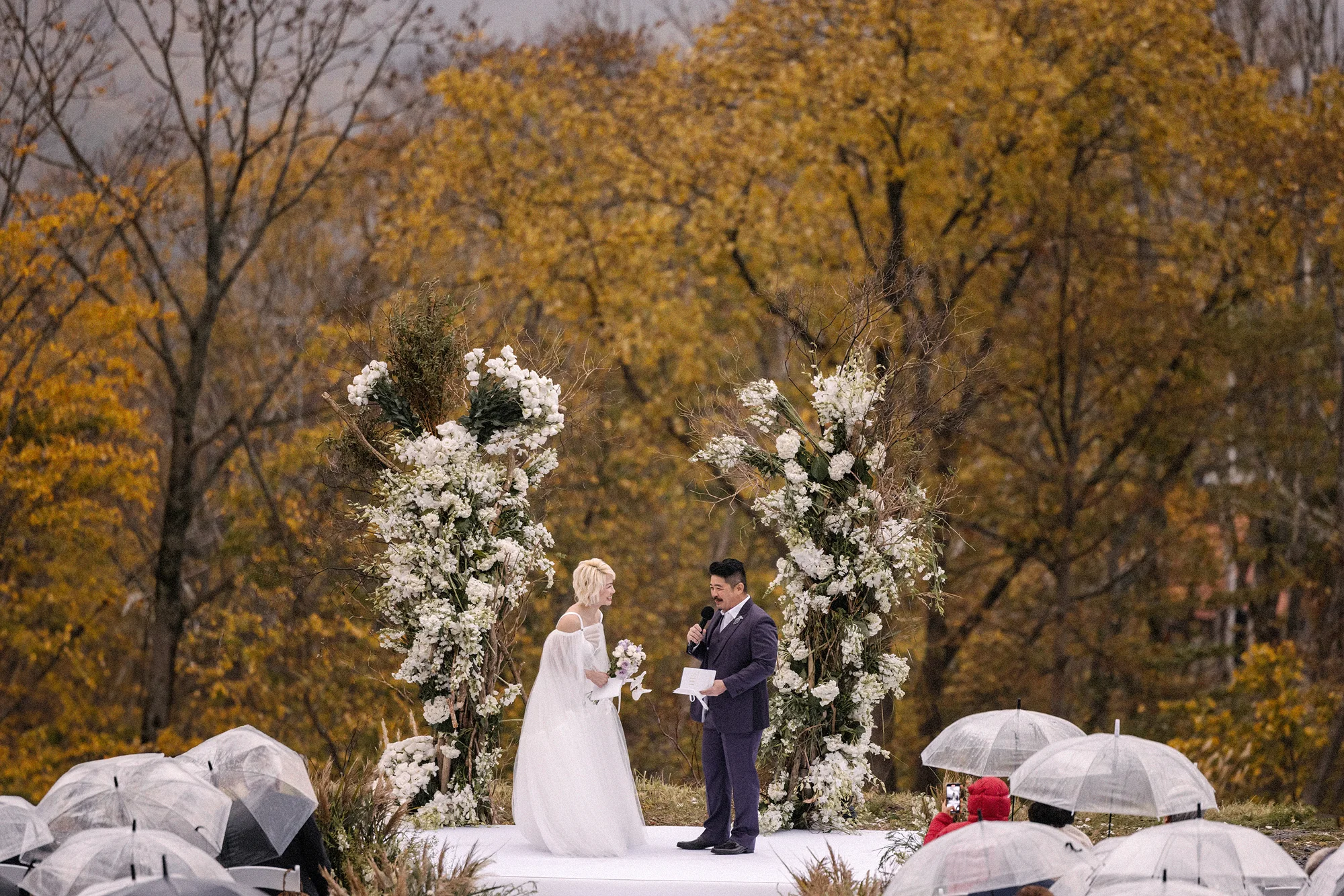 Guests under clear umbrellas watch the couple at the top of an aisle between white flowers and autumn trees.