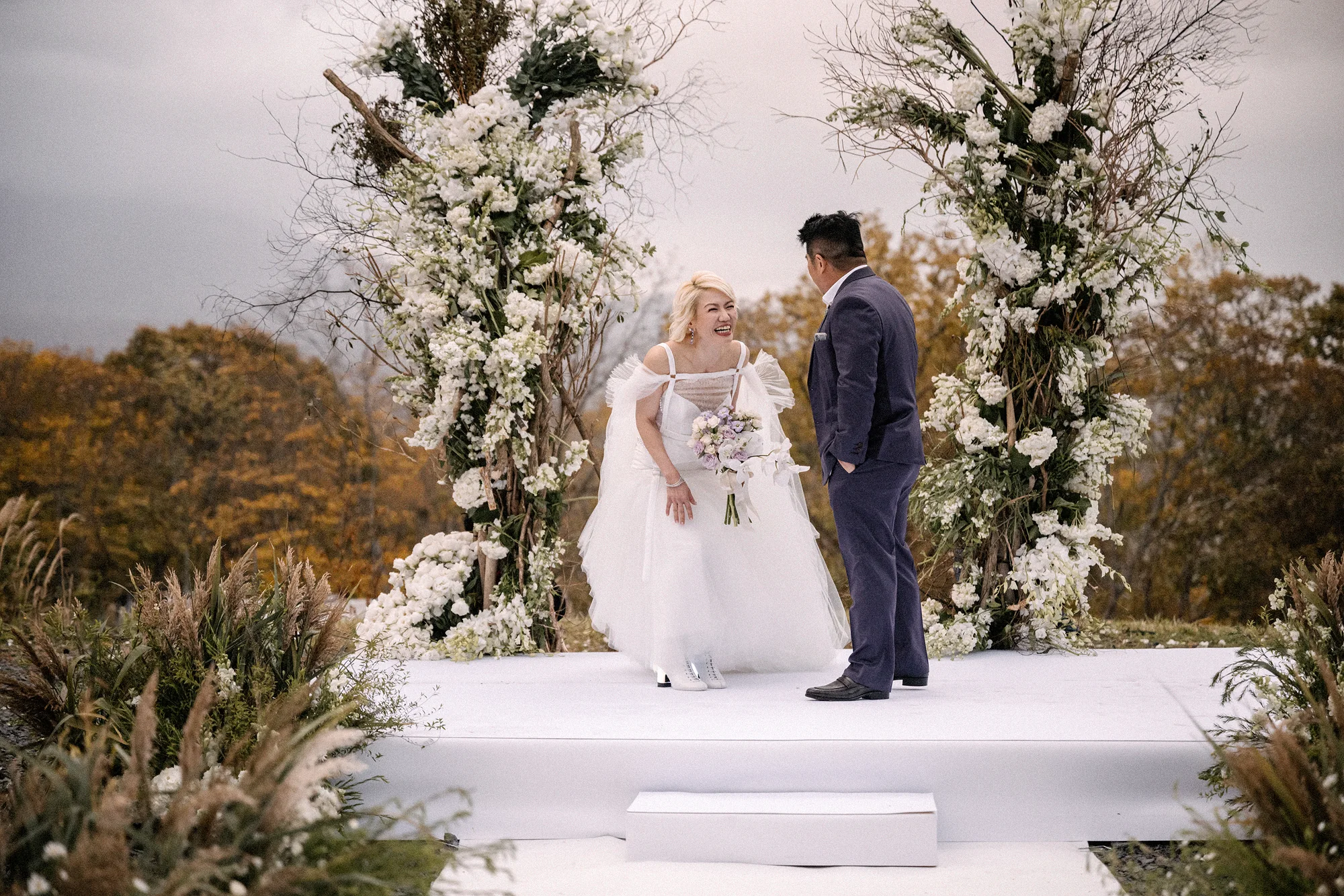 The bride and groom between tall columns of white flowers at an outdoor ceremony, autumn trees behind.