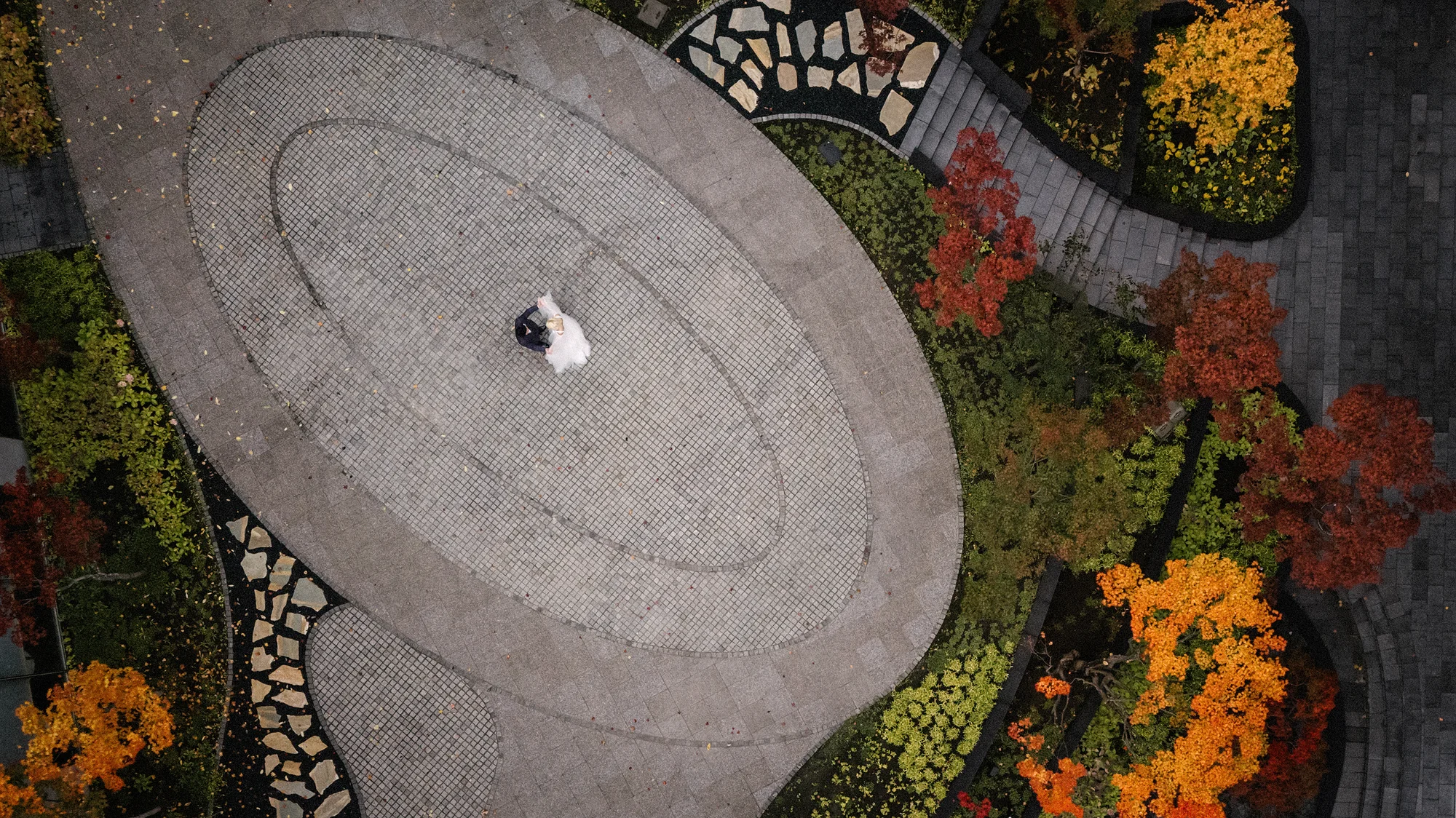 From above, the couple stand in an oval stone courtyard ringed with autumn planting in Niseko.