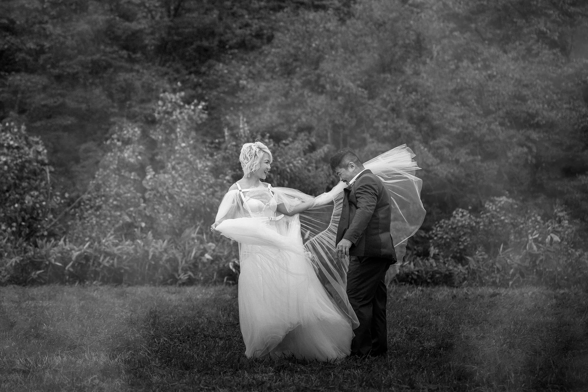 In black and white, the wind wraps the bride's cape around the groom in an open field.