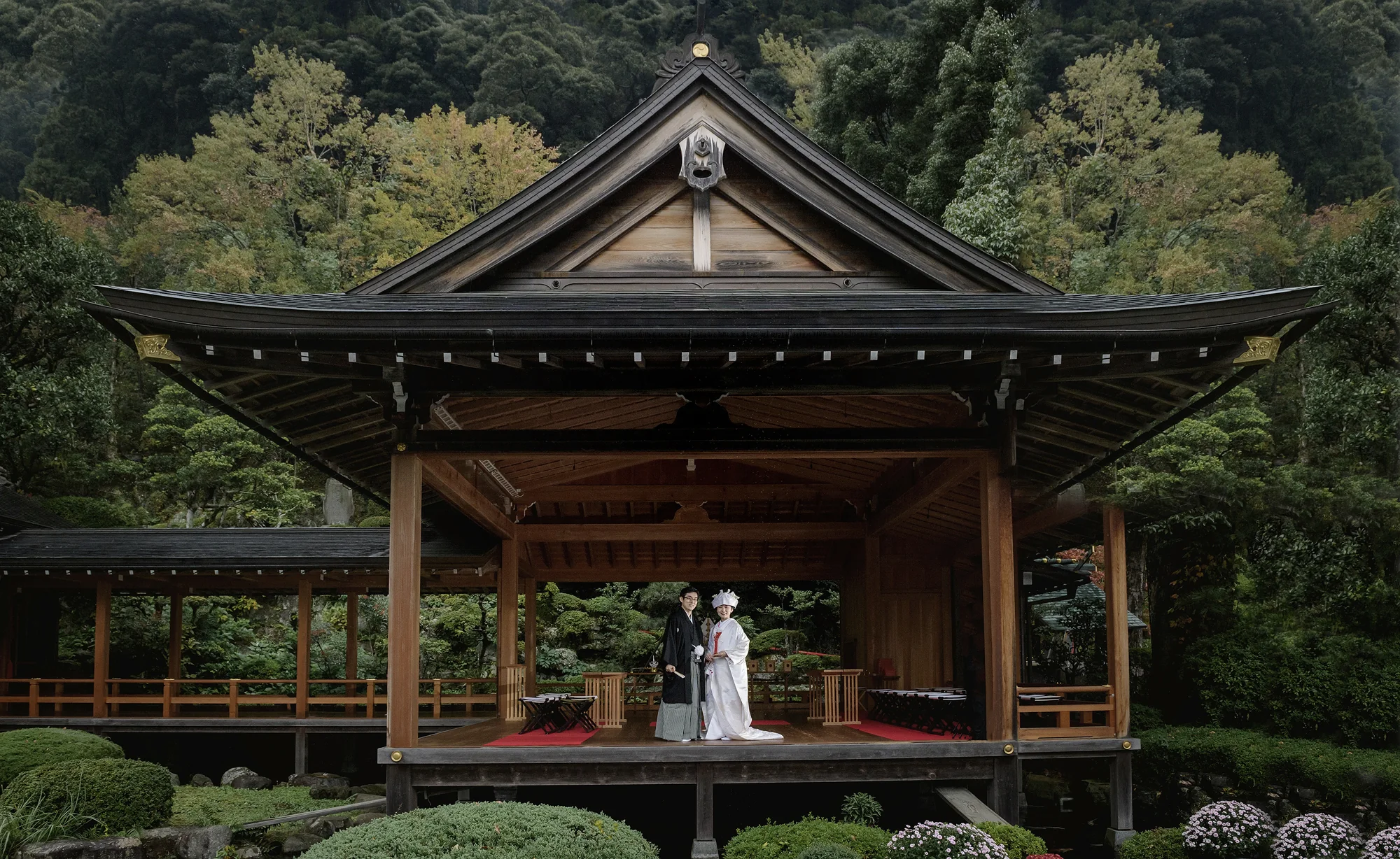 A couple stand on an open Noh stage in a green garden, the wooden roof sweeping above them.