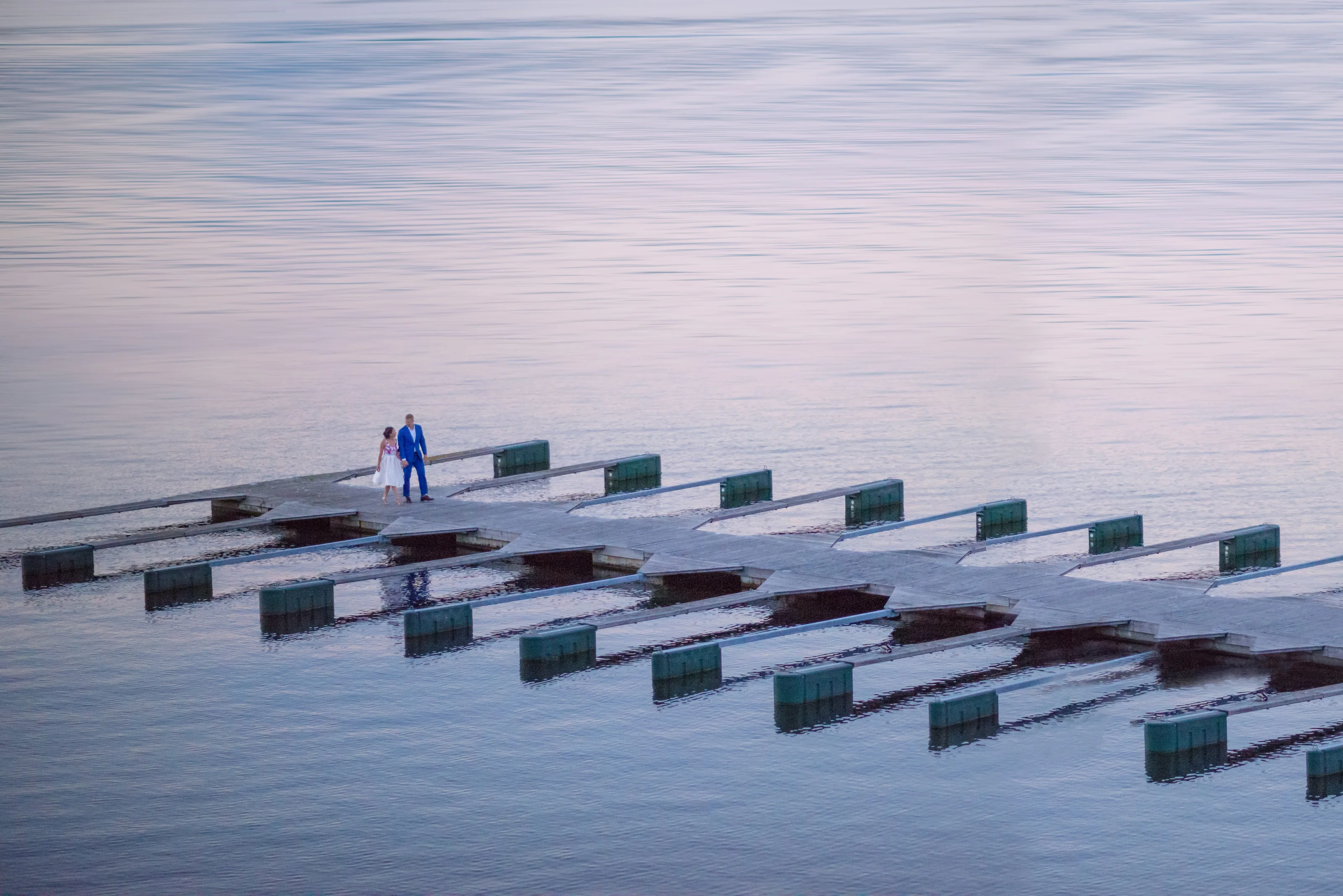 A couple stand small on a jetty over still water under the pale light of the midnight sun in Norway.