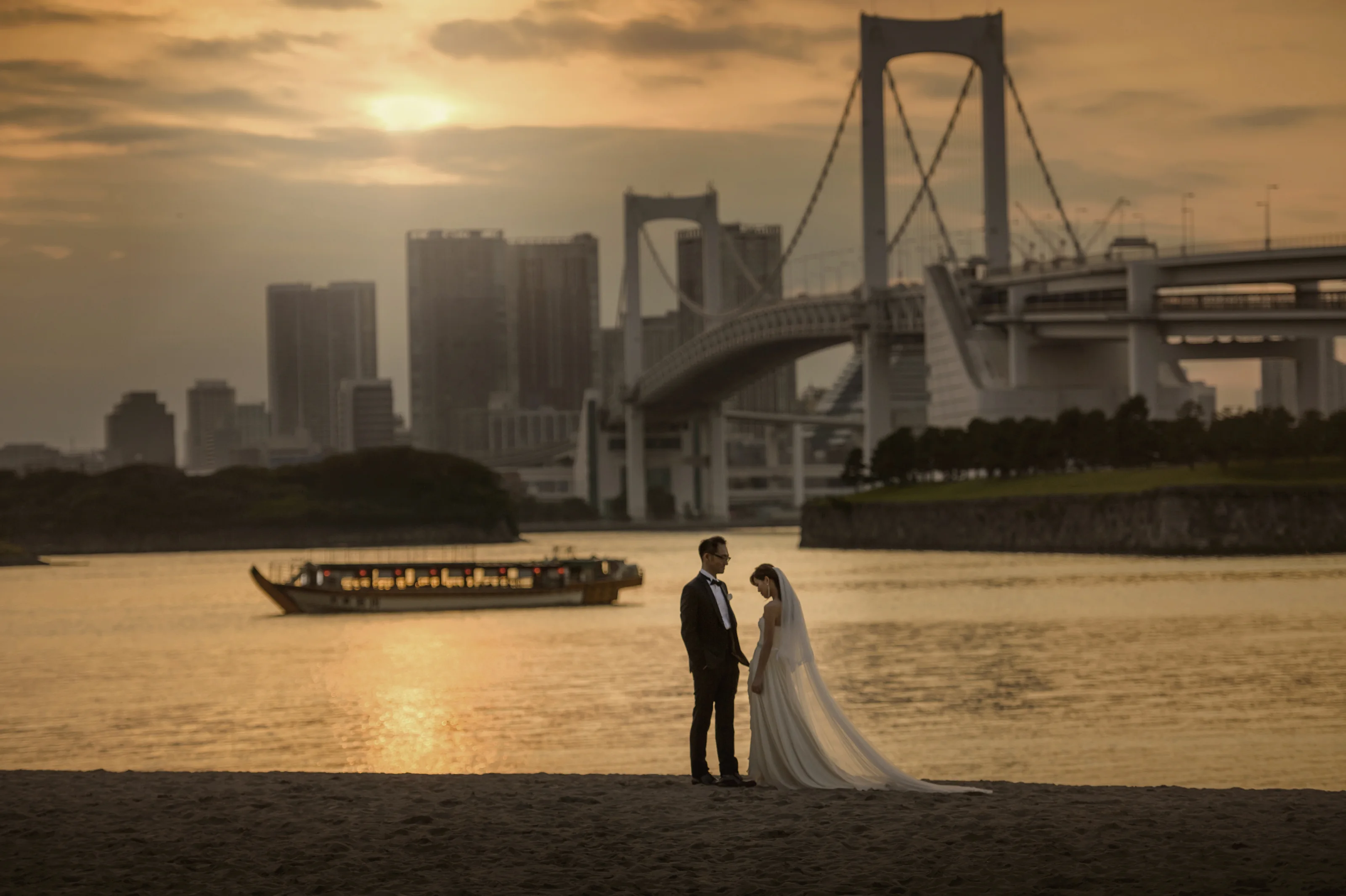 The couple stand on the shore at Odaiba as a golden sunset lights Tokyo Bay, the Rainbow Bridge and a yakatabune boat behind them.