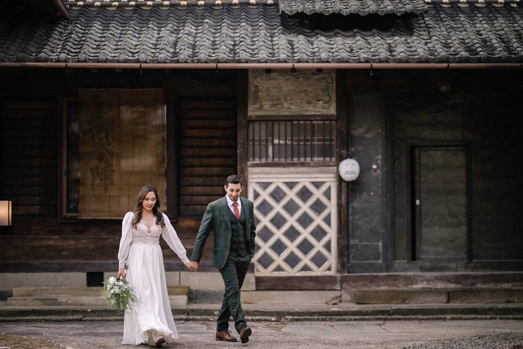 The couple walk hand in hand past the timber front of an old Japanese farmhouse.