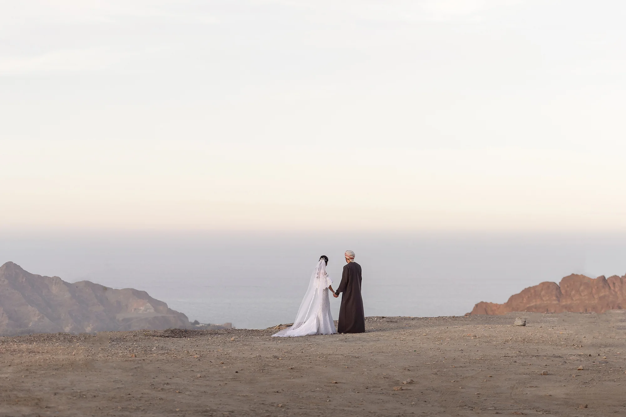 The couple stand hand in hand at the edge of a clifftop above the sea in soft evening light in Oman.