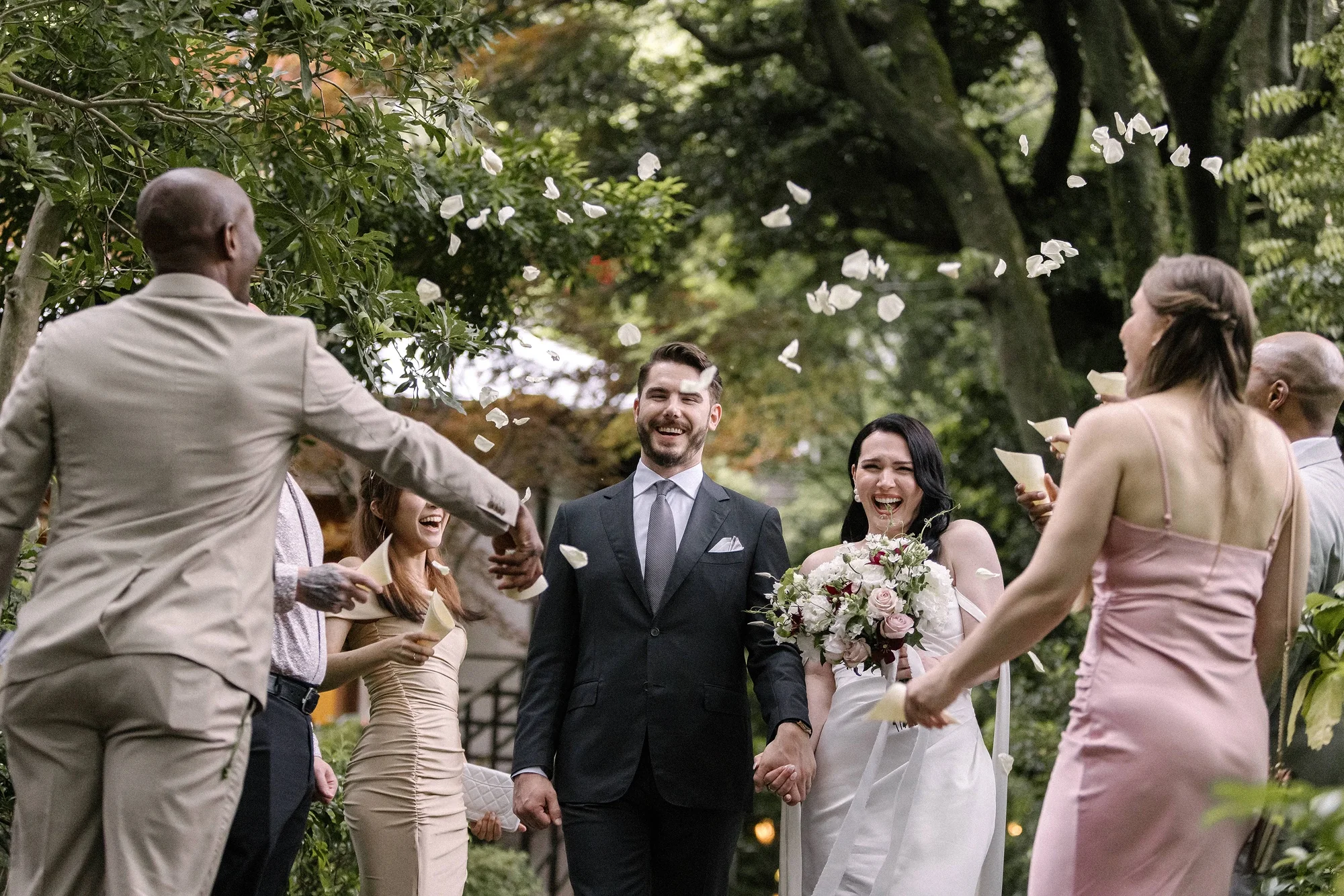 Guests throw petals as the couple walk back down the aisle in a garden, laughing.