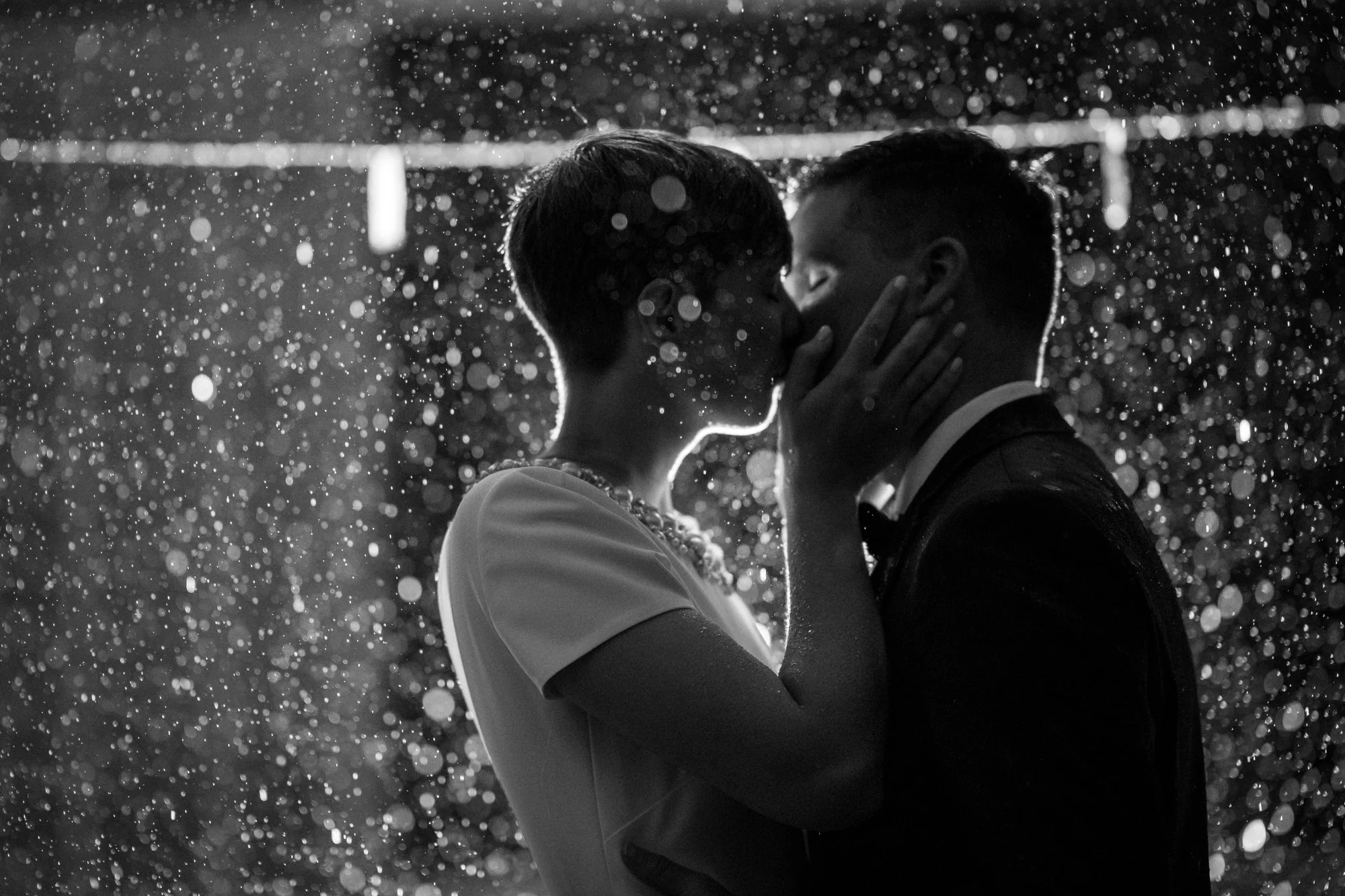 In black and white, a couple kiss in the rain, the drops lit around them.