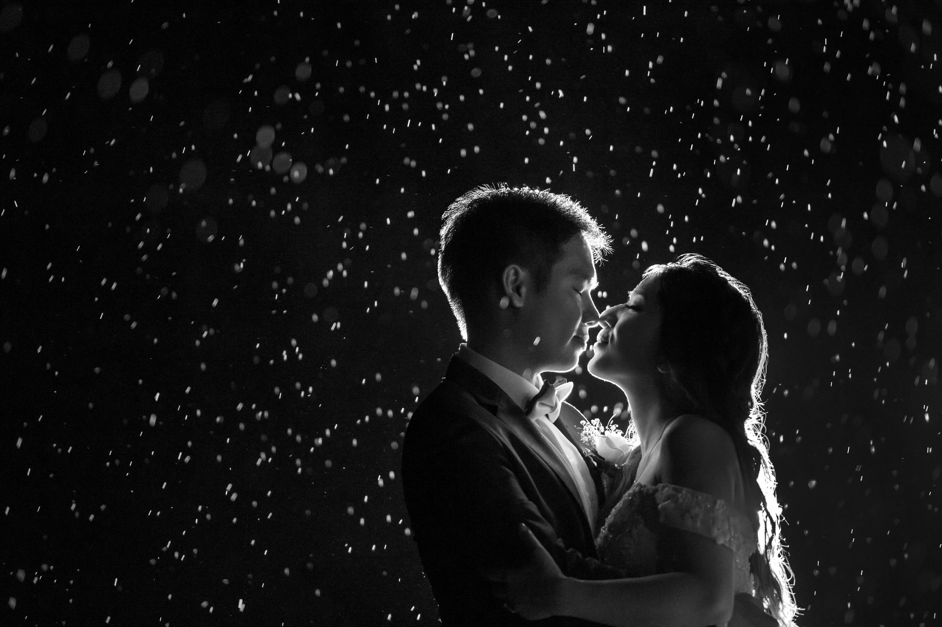 In black and white, the couple lean in for a kiss as rain falls through the light around them.