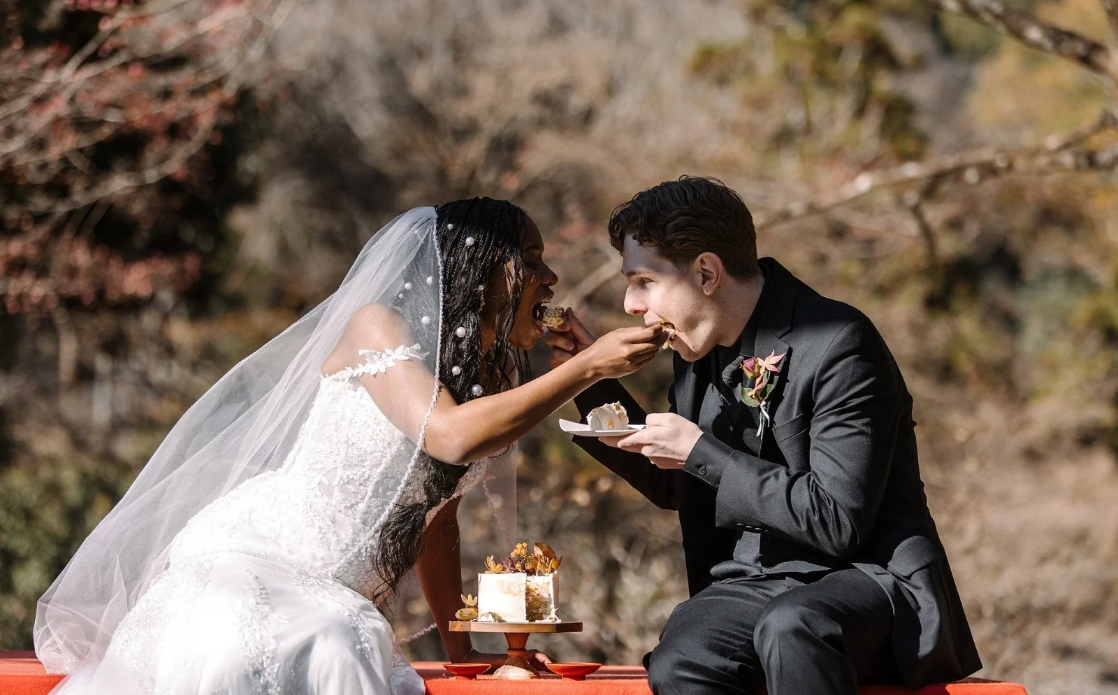 The bride feeds the groom a bite of cake as they sit on a red bench among autumn trees.