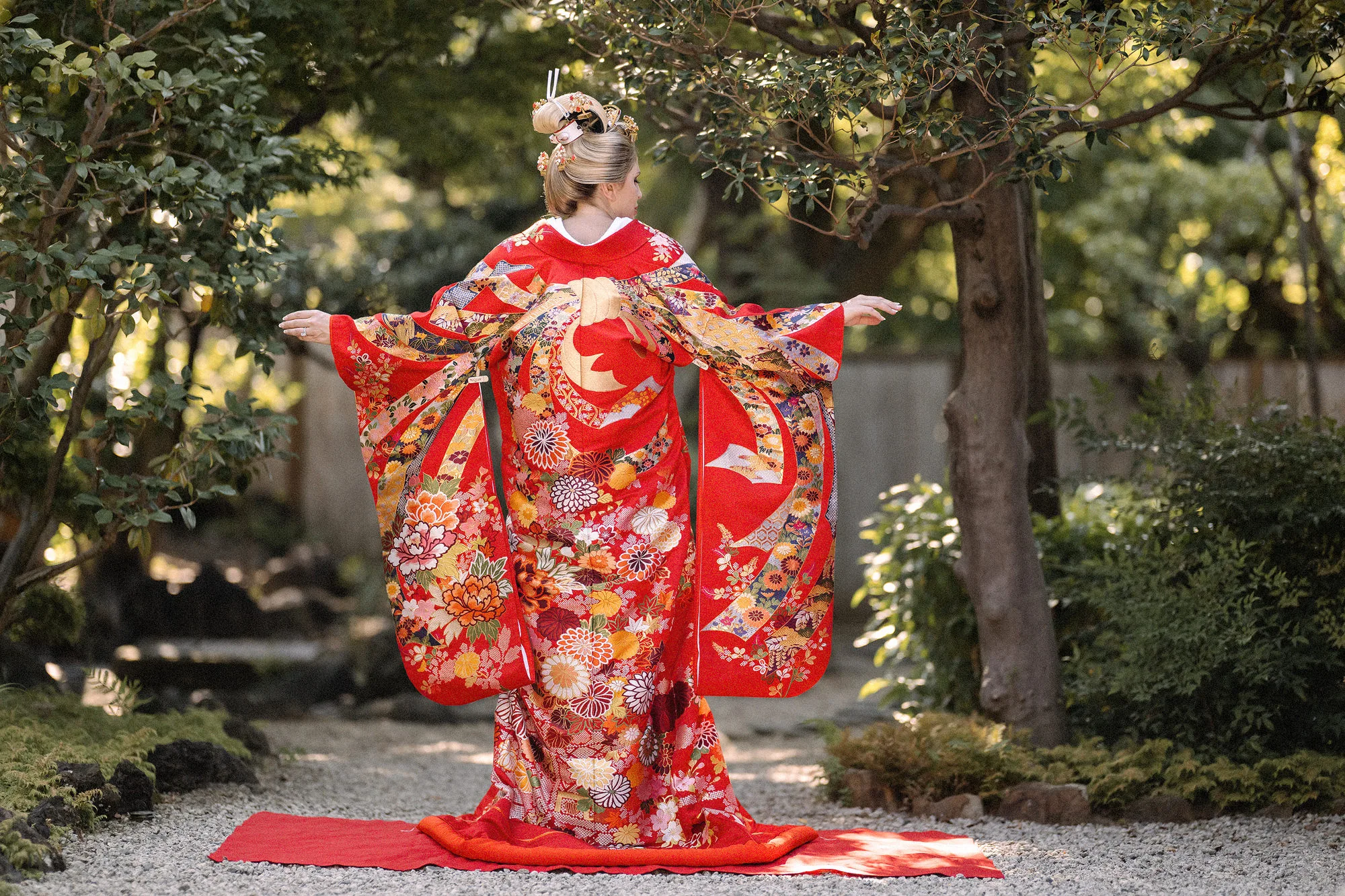 Bride in a red iro-uchikake wedding kimono and nihongami, arms open to show the back of the robe, on a gravel path in a Tokyo garden